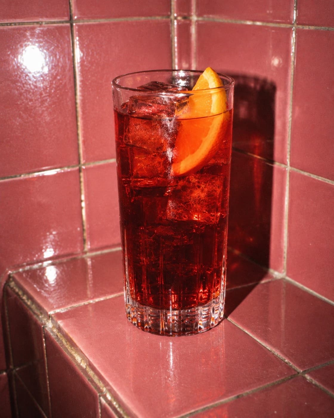 A bright red Americano over ice with an orange slice in a short glass on a pale speckled counter
