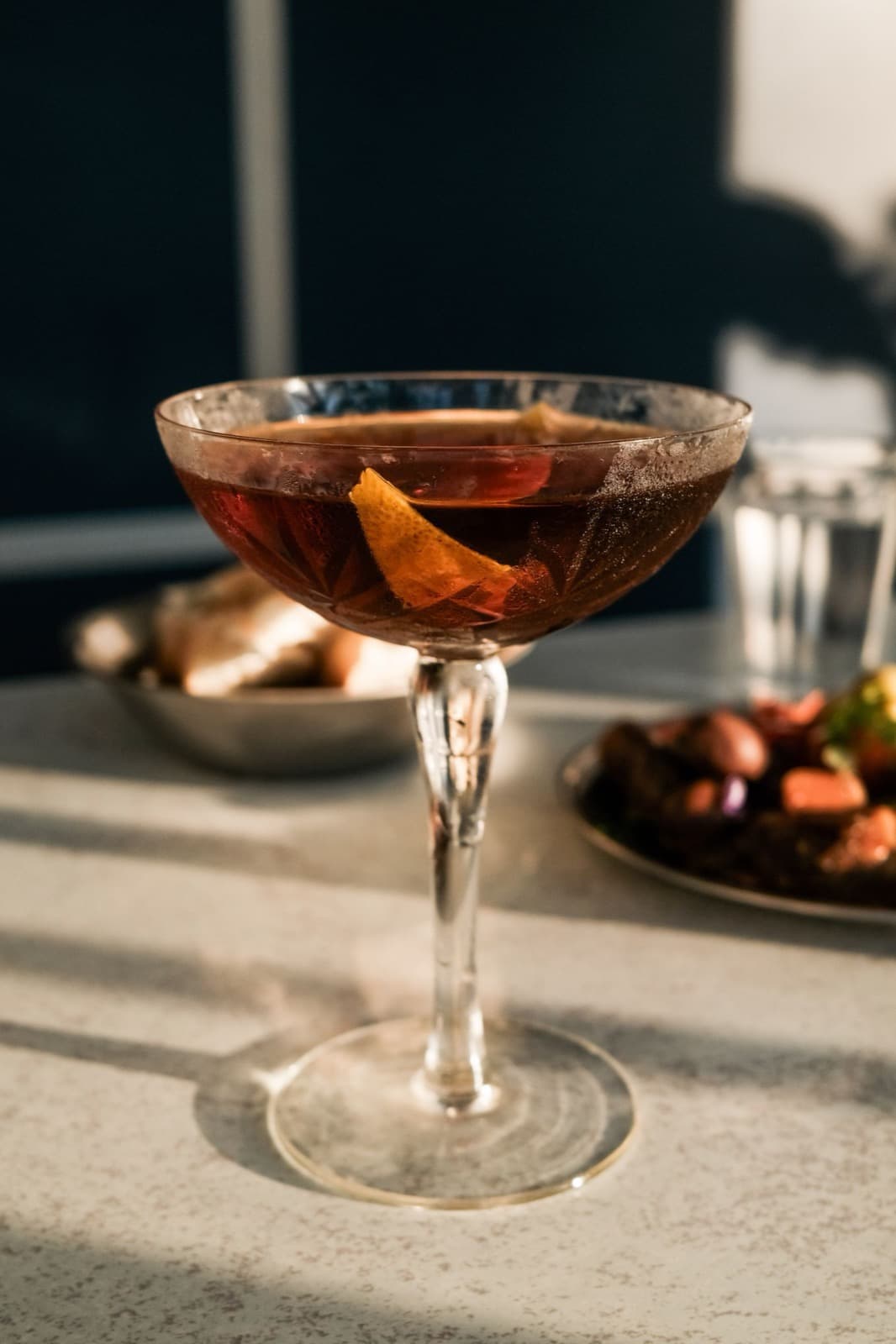 A dark mahogany drink in a stemmed coupe with an orange peel, on a sunlit stone table beside small plates