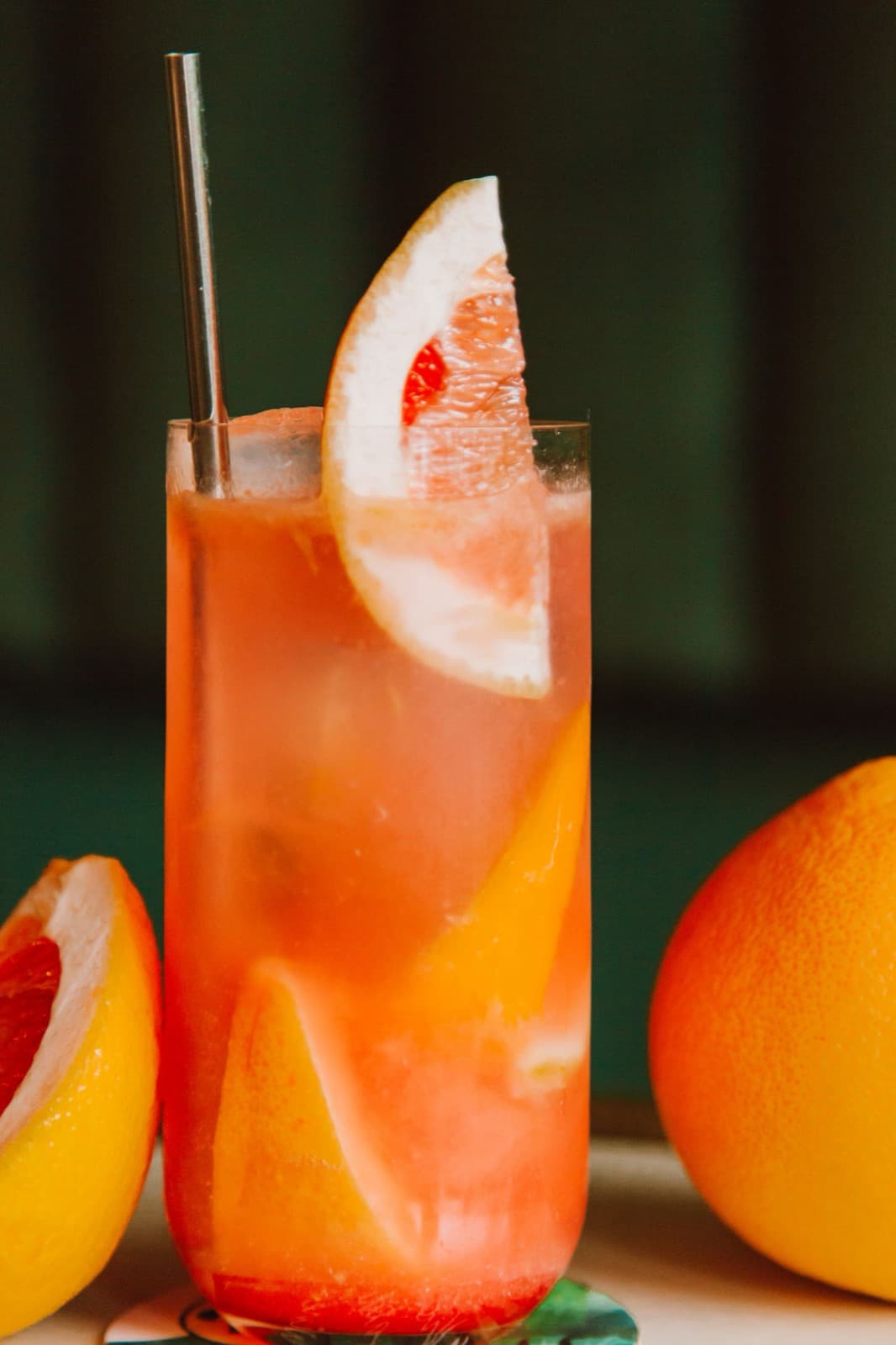 A tall pink-orange grapefruit drink on ice with a grapefruit slice and a metal straw, beside halved grapefruit