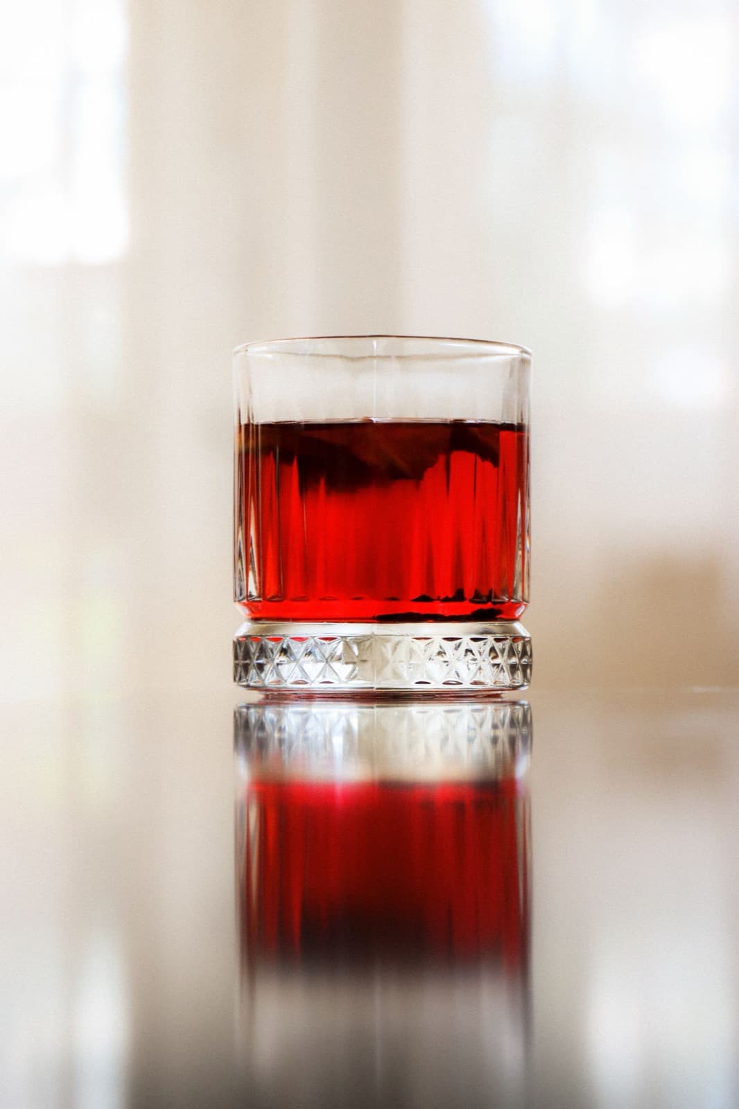 A ruby red drink in a short ribbed tumbler on a glossy surface, against soft pale window light