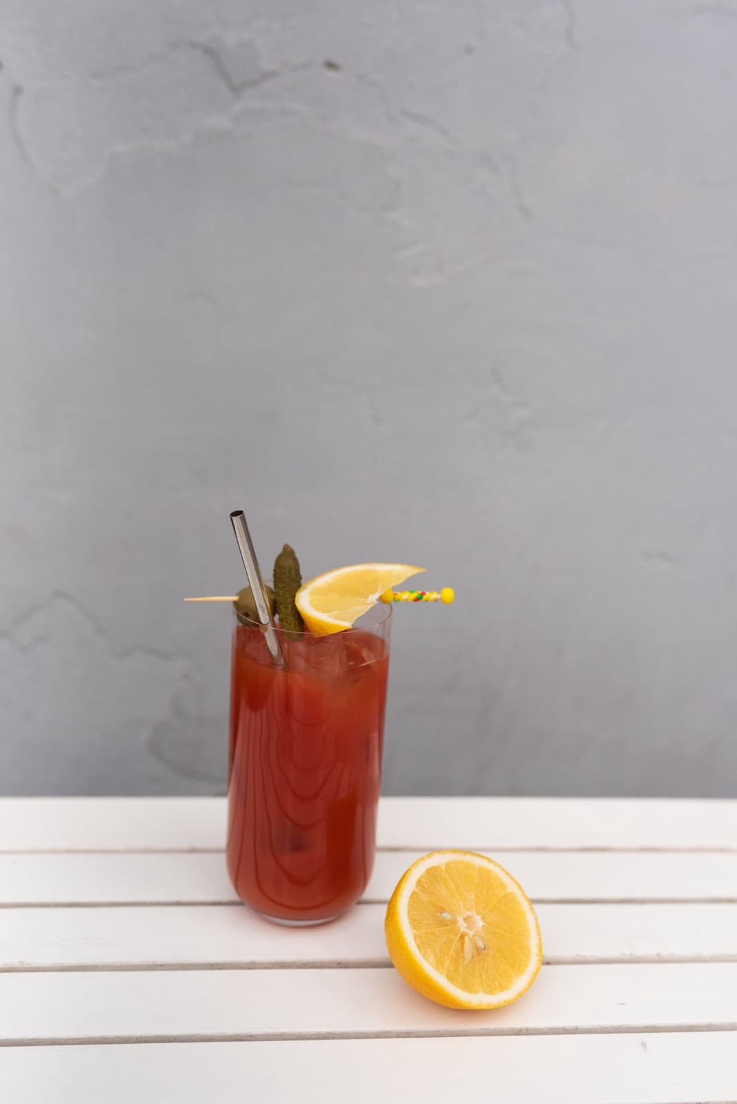 A Bloody Mary in a tall glass with a lemon and pickle garnish on a pick, beside half a lemon on a white slatted table