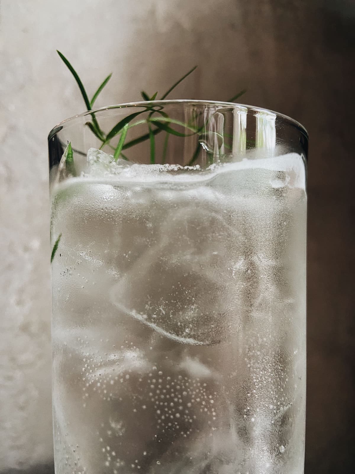 A tall glass of sparkling tonic over ice with sprigs of green herbs, against a soft pale wall