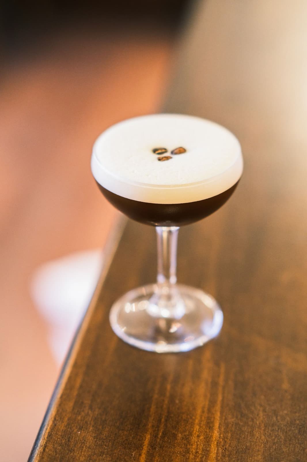 An espresso martini in a coupe with a thick crema and three coffee beans, on the warm wooden corner of a table