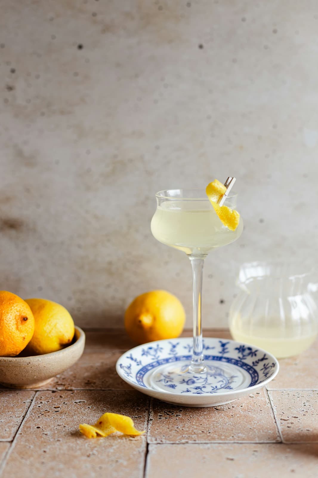 A pale citrus daiquiri in a coupe with a twist of peel, on a blue-and-white saucer beside a bowl of lemons on warm stone
