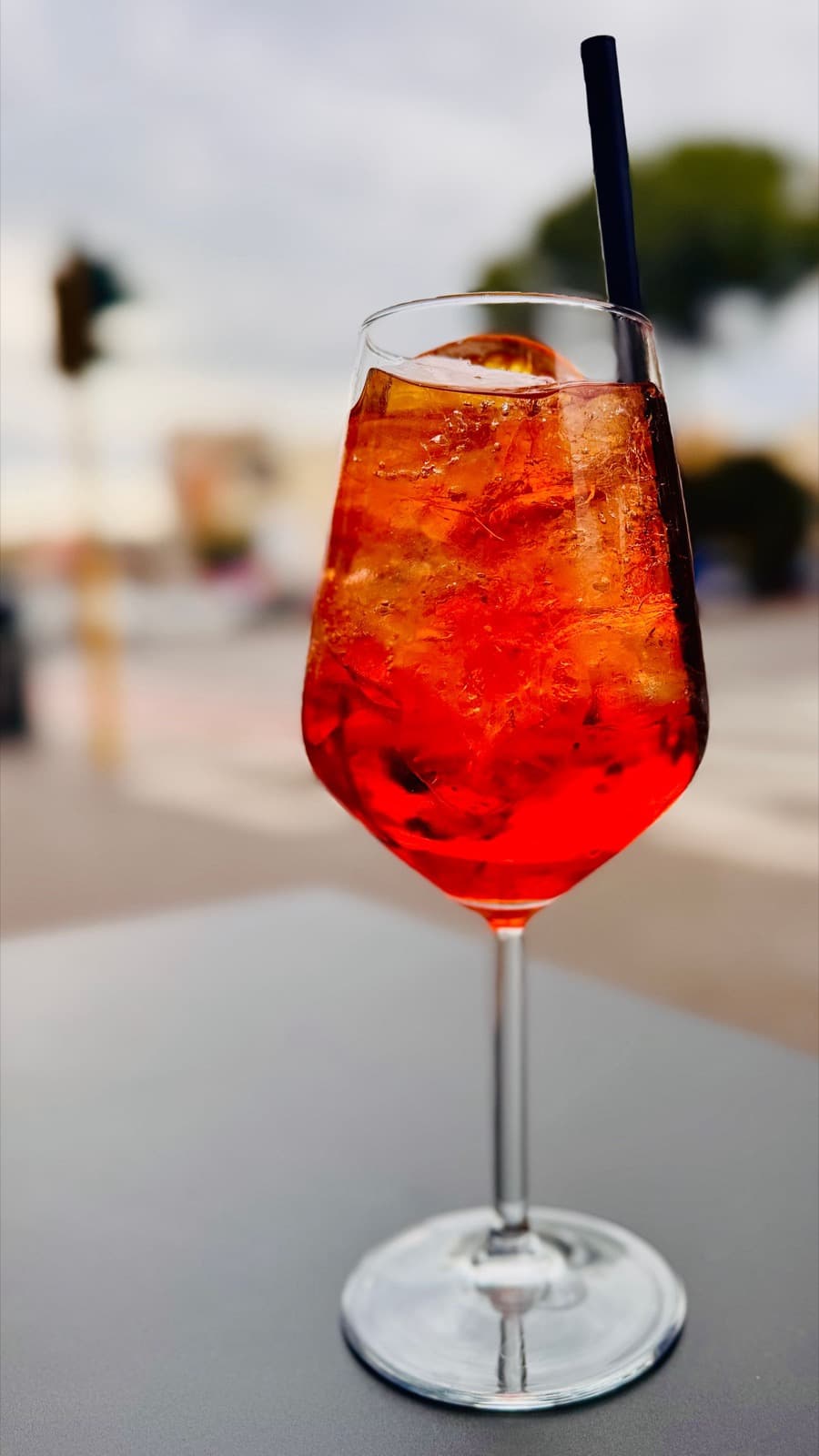 A bright orange spritz packed with ice in a stemmed wine glass with a black straw, on a pale outdoor table