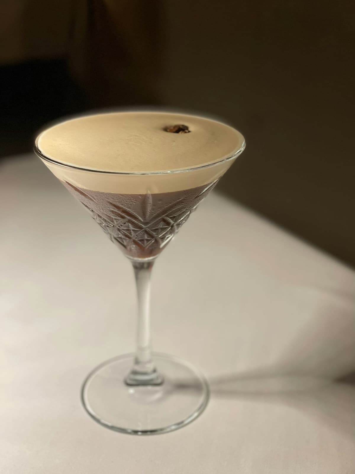 An espresso martini with a pale crema and a single coffee bean in a cut-glass martini glass on a white tablecloth