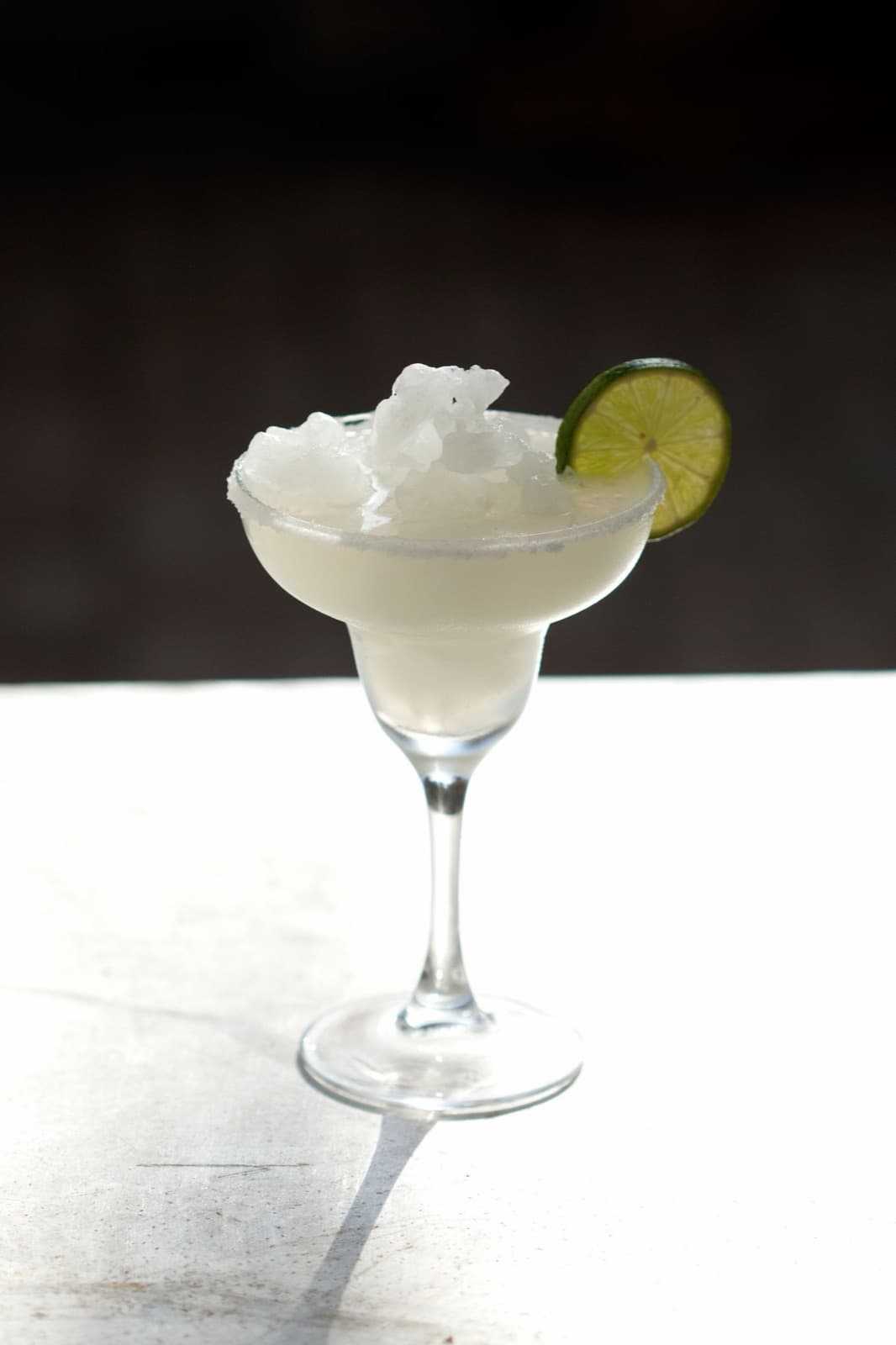 A pale frozen margarita heaped with slush in a stemmed margarita glass with a lime wheel, on a bright white table