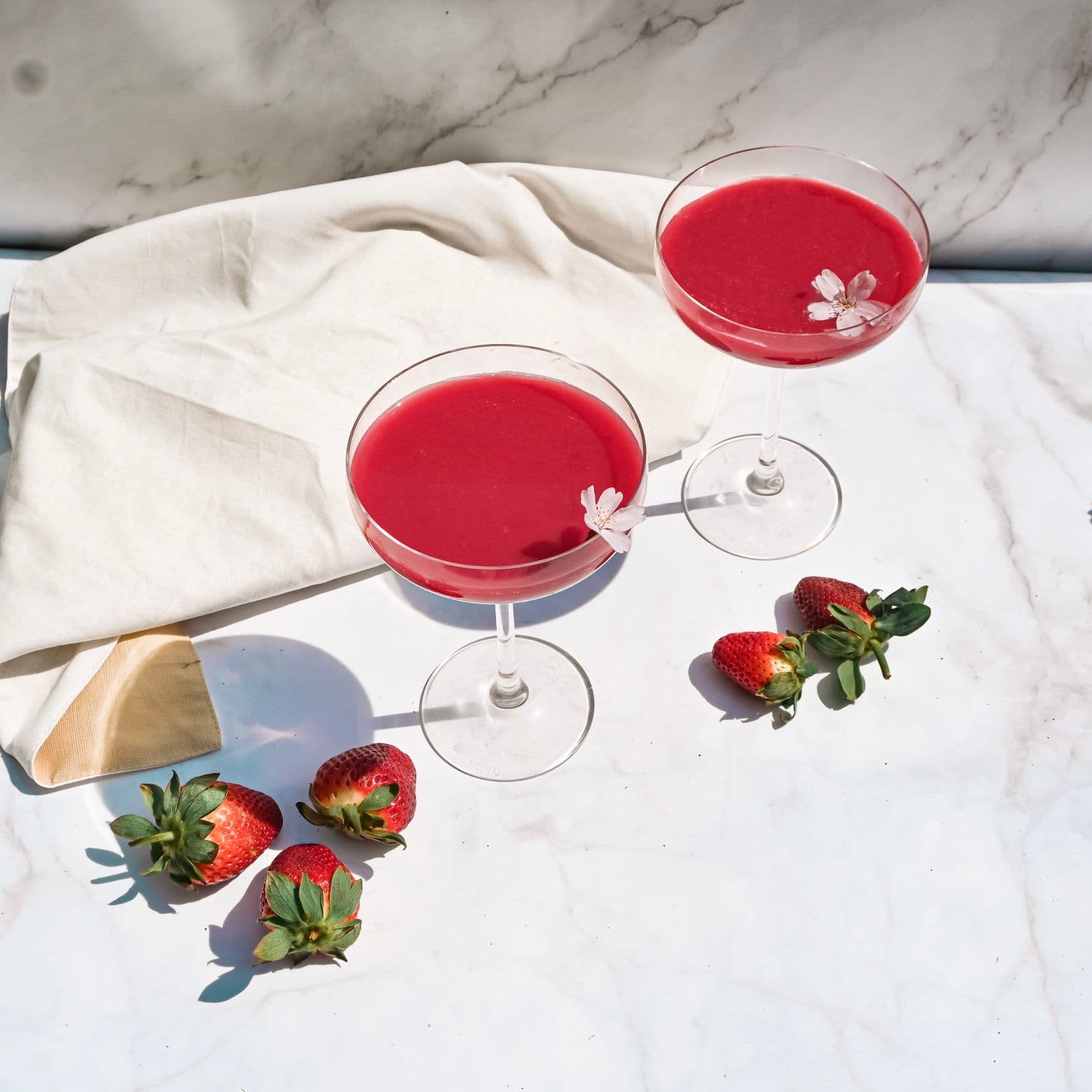 Two strawberry daiquiris in coupes with small flower garnishes, fresh strawberries scattered on white marble in sunlight