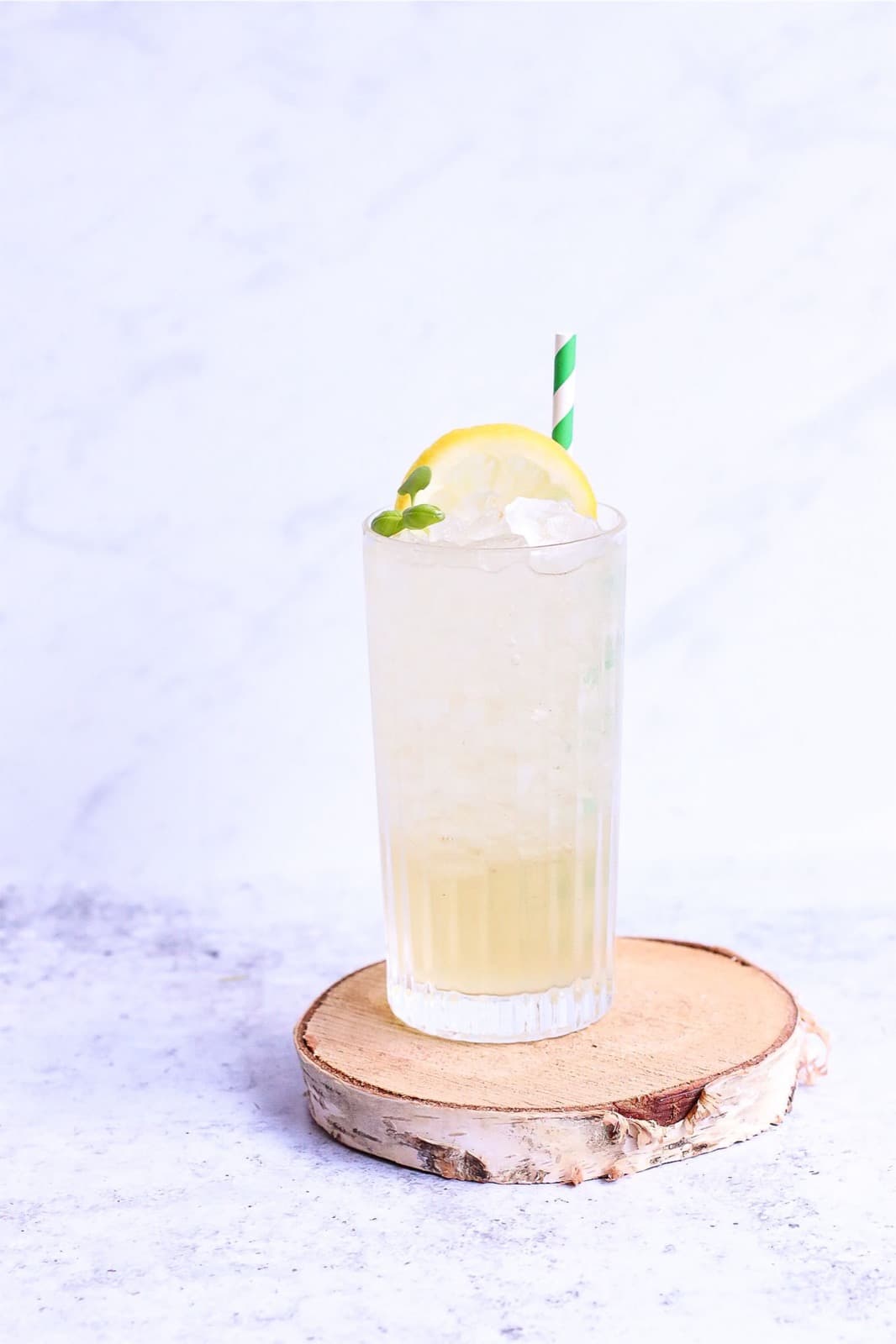 A pale, fizzy gin fizz over ice in a tall ribbed glass with a lemon wheel, mint and a striped straw, on a birch slice against white marble