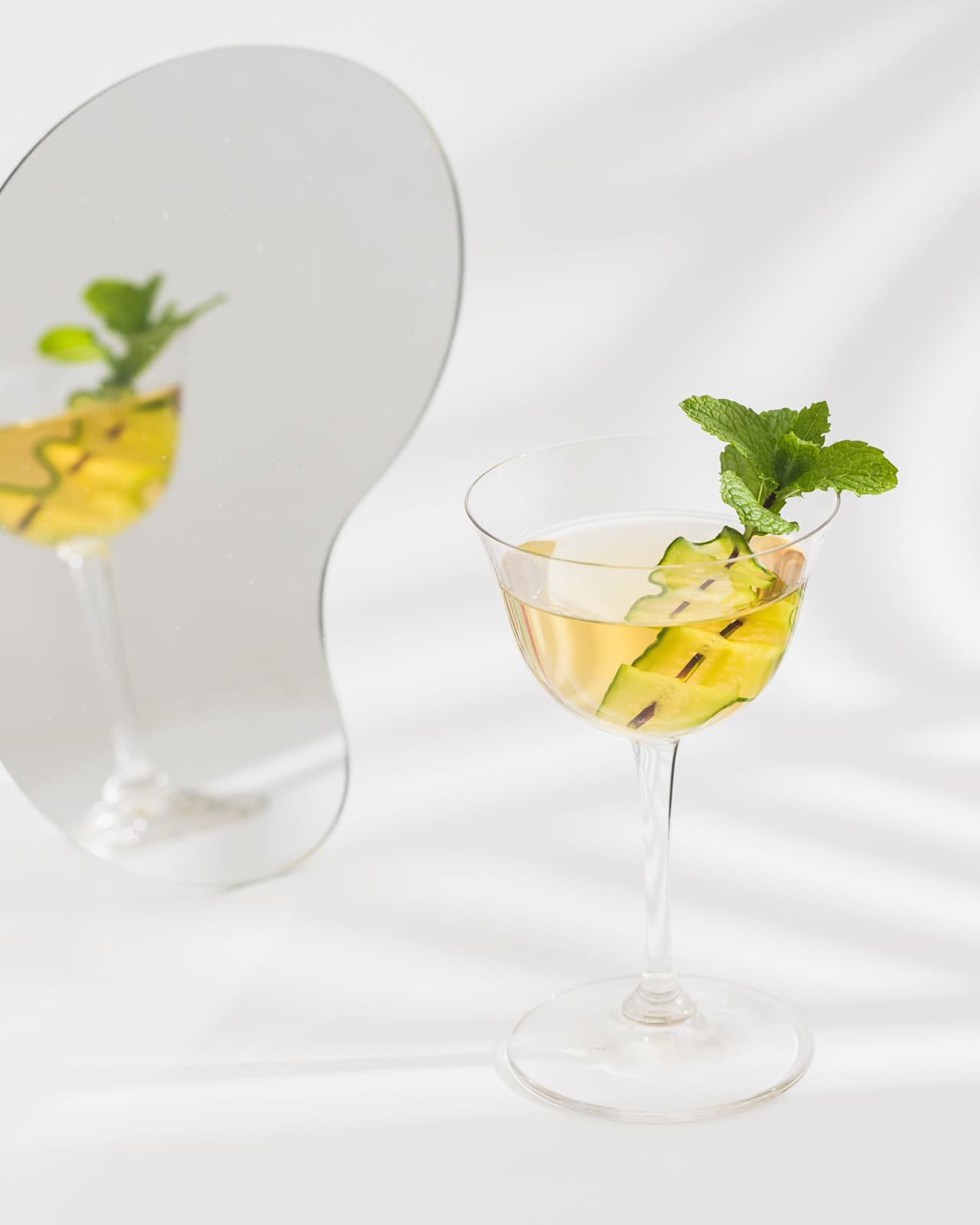 A pale green-gold drink in a stemmed glass with a mint sprig and cucumber ribbons, reflected in a curved mirror on a white table