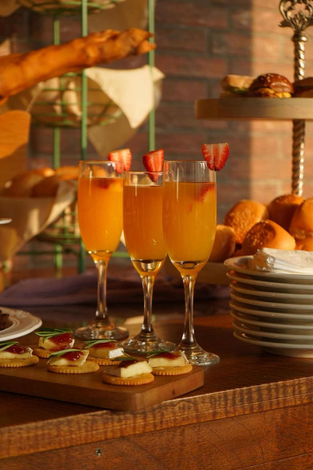 Three mimosas in stemmed glasses topped with strawberries, beside a brunch spread of pastries and canapés in warm light