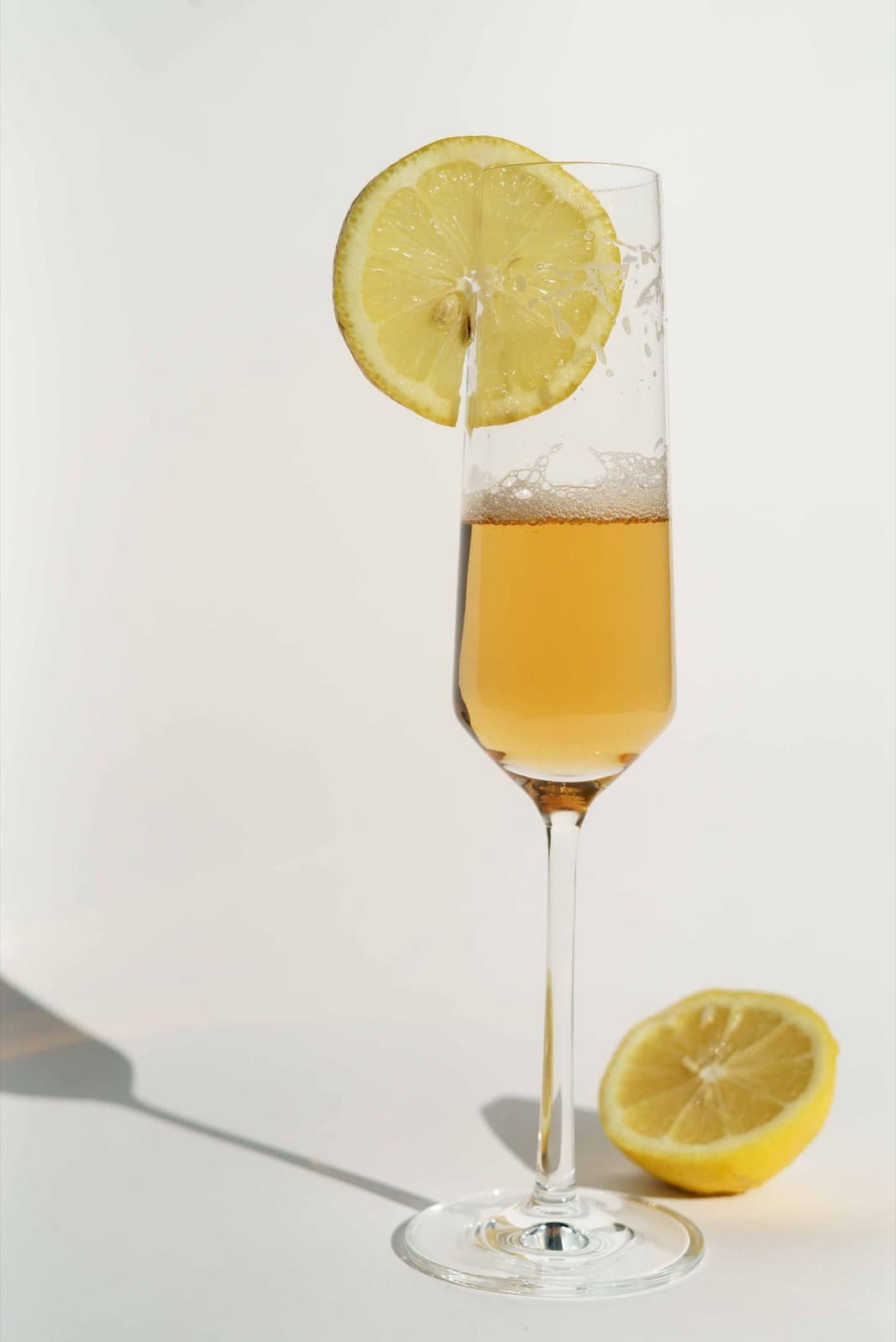 A sparkling citrus drink in a champagne flute with a lemon wheel on the rim, beside half a lemon on a white surface