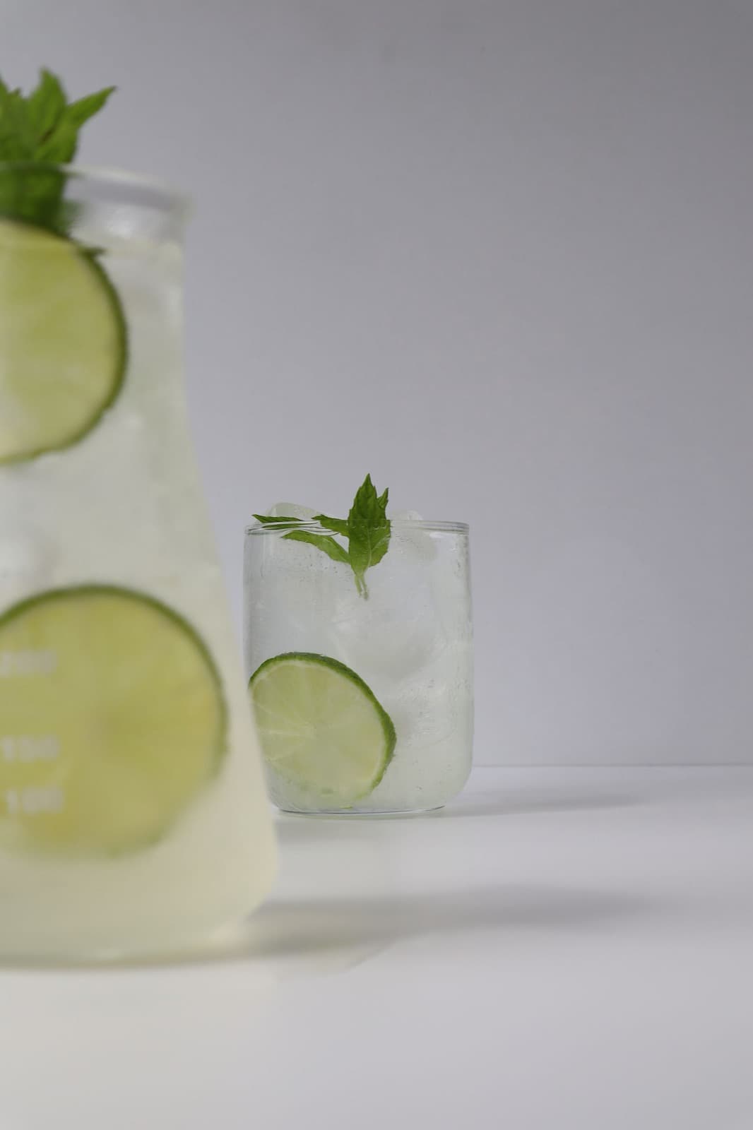 A glass pitcher of lime and mint mojito beside a short glass with a lime wheel and mint sprig, on a pale grey surface