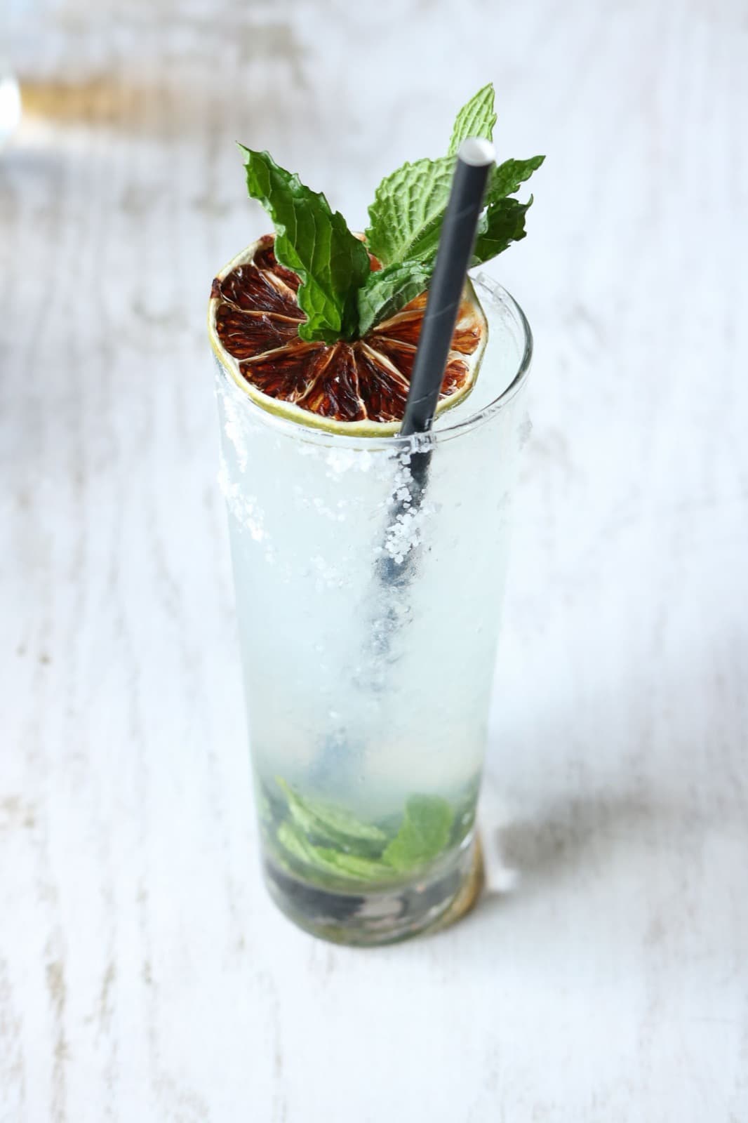 A tall mojito topped with fresh mint and a dried lime wheel on a white wooden table