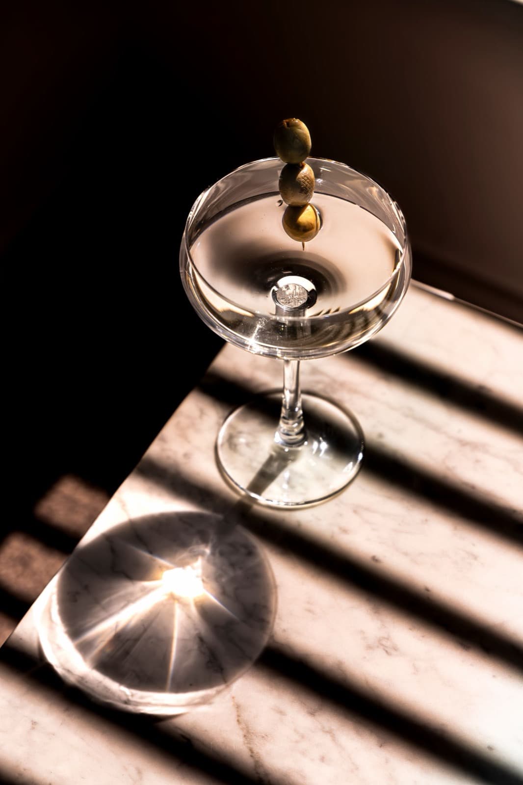 A dirty martini in a coupe with a pick of green olives, on a marble table striped with window-blind shadows