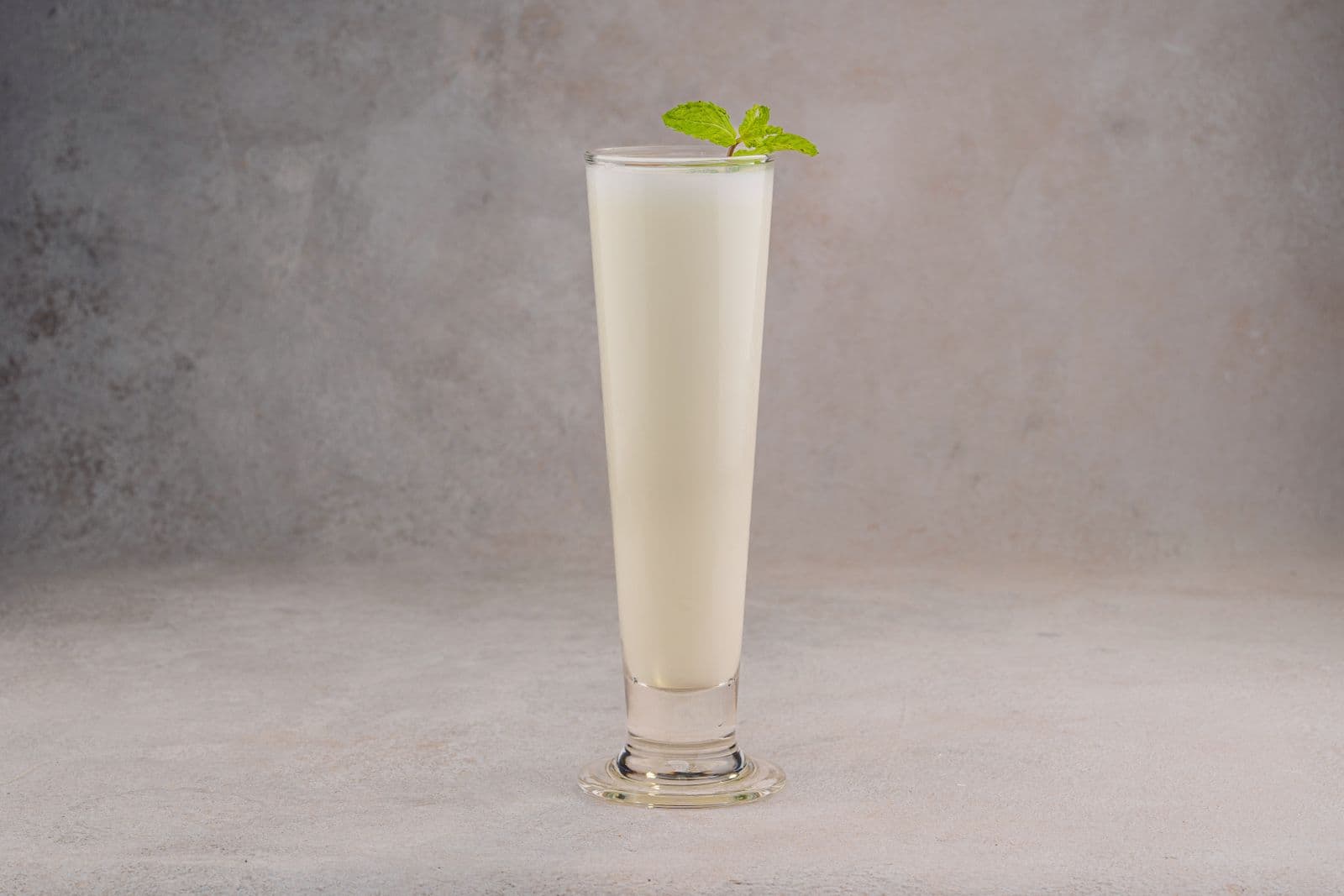A tall, creamy white fizz in a footed glass topped with a small mint sprig, against a soft grey backdrop