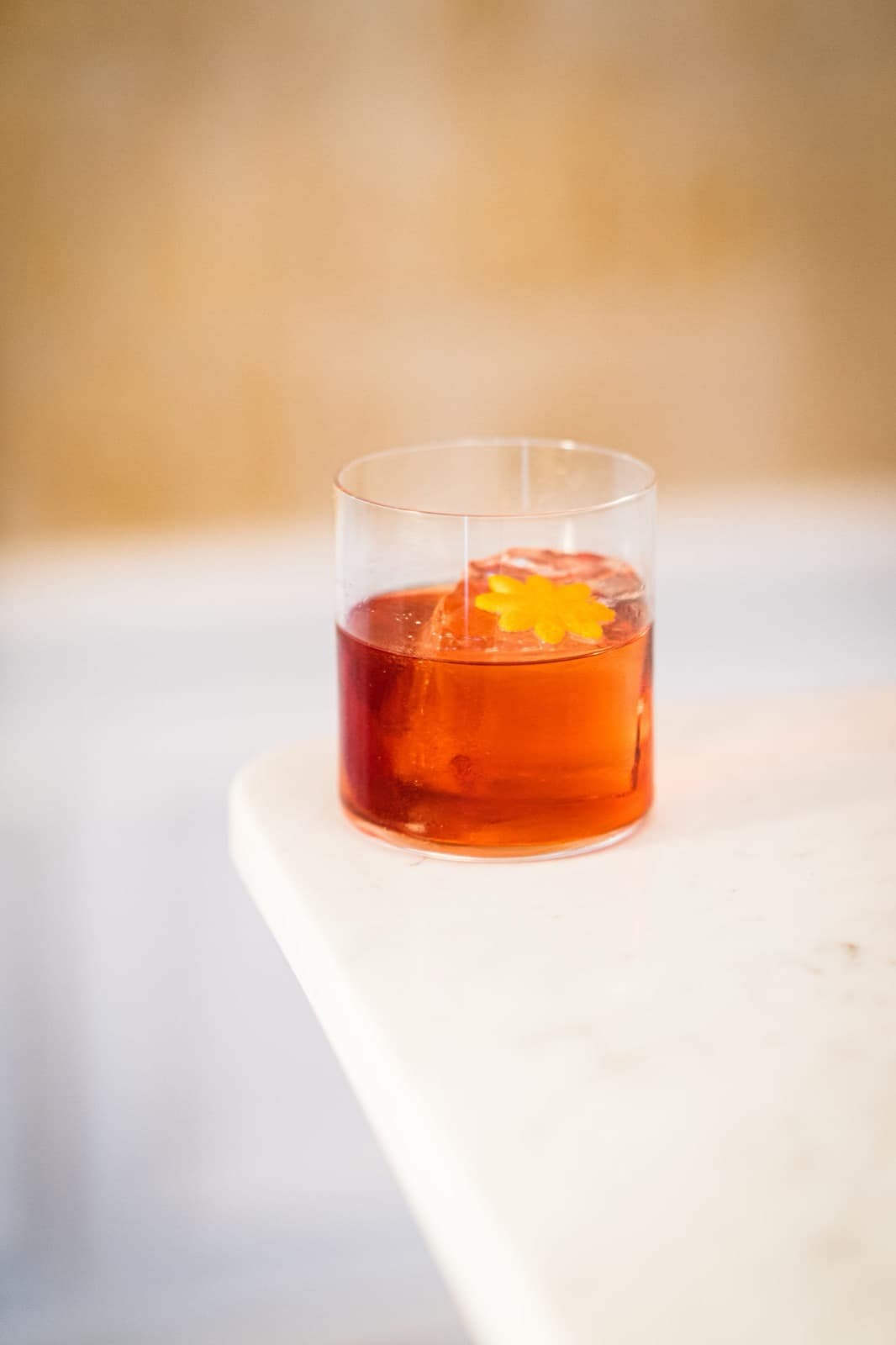 A negroni on ice with an orange twist in a short tumbler, on a pale marble ledge against a warm cream wall