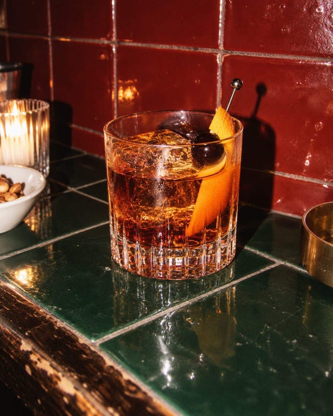 An old fashioned over a large ice cube with an orange peel on a pick, on a cream stone surface