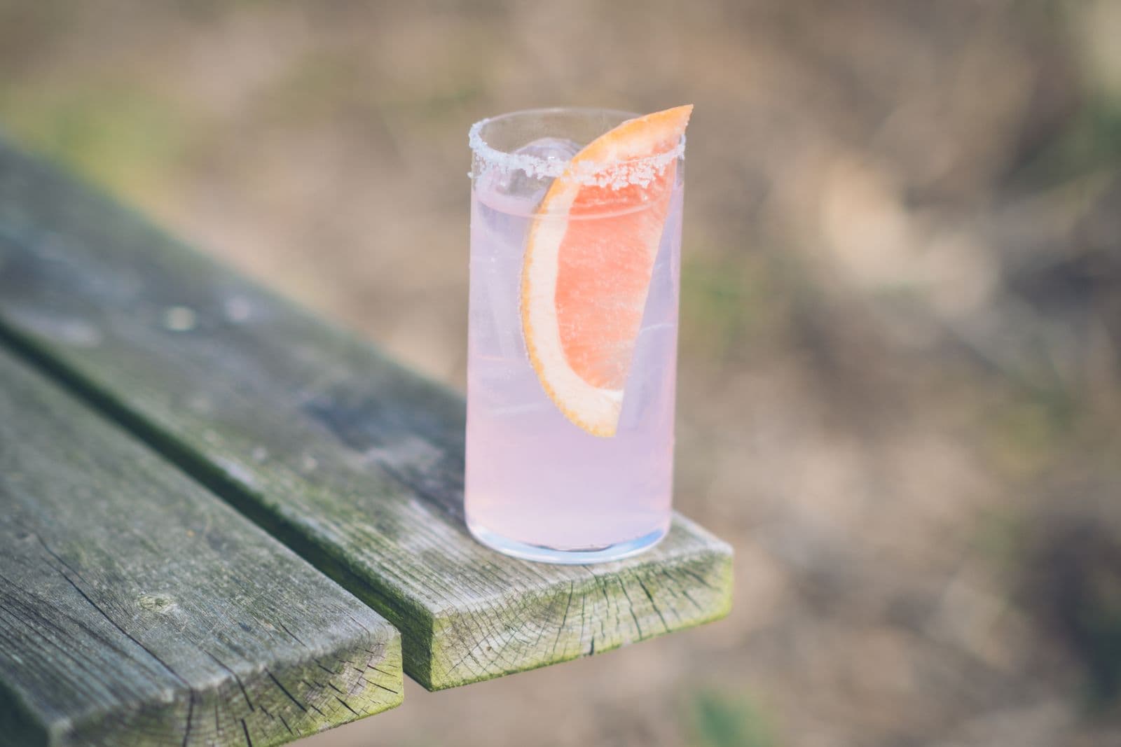 A pale pink grapefruit drink with a salted rim and a grapefruit wedge in a tall glass, on a weathered wooden bench