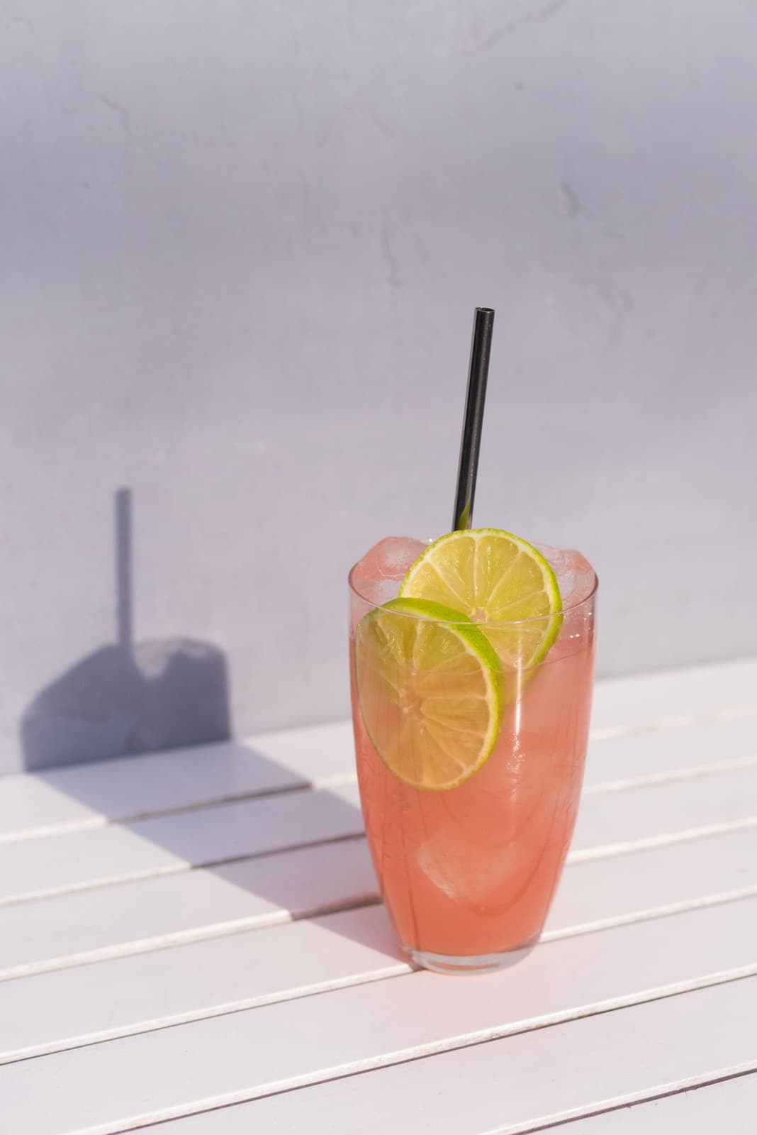 A pink grapefruit highball with lime wheels and a straw on a white slatted table