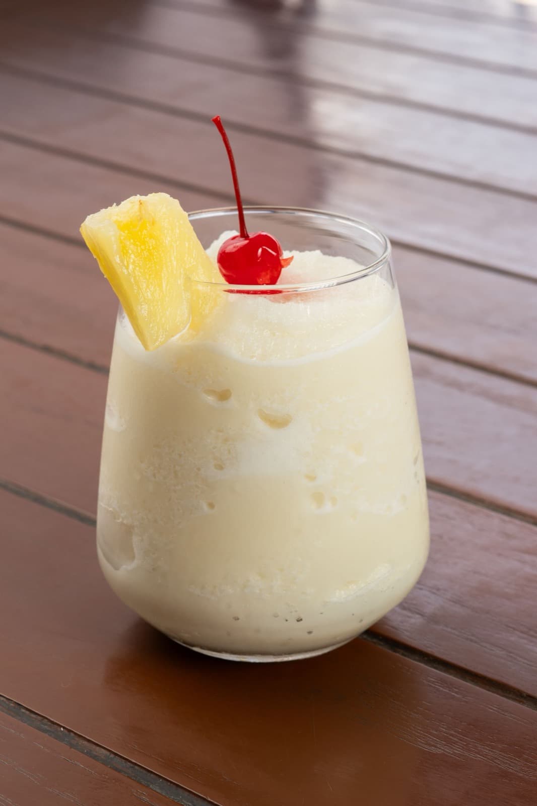 A creamy piña colada over ice in a tumbler with a pineapple wedge and cherry, on a warm wooden table