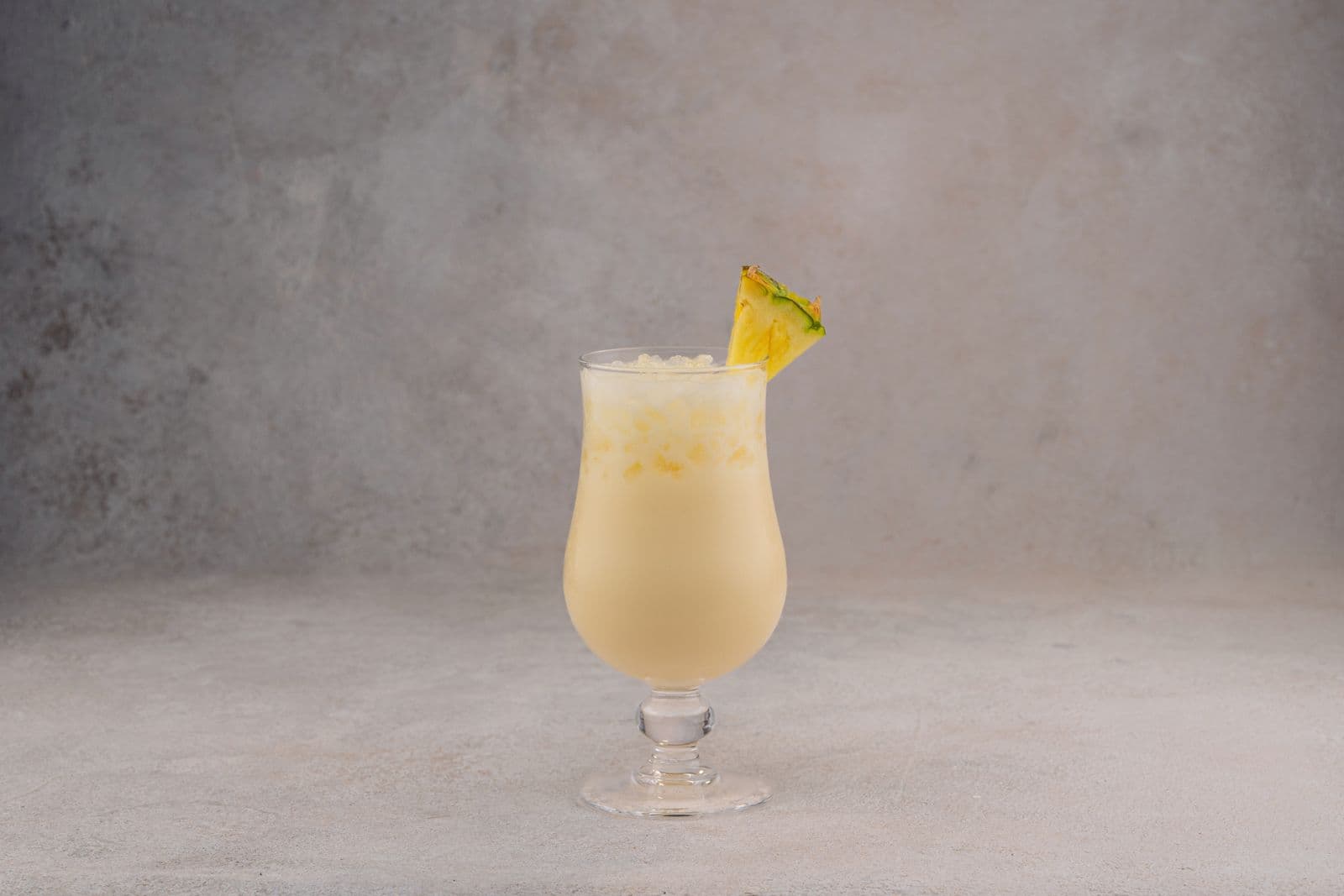 A creamy piña colada in a hurricane glass with a pineapple wedge, against a pale grey textured background