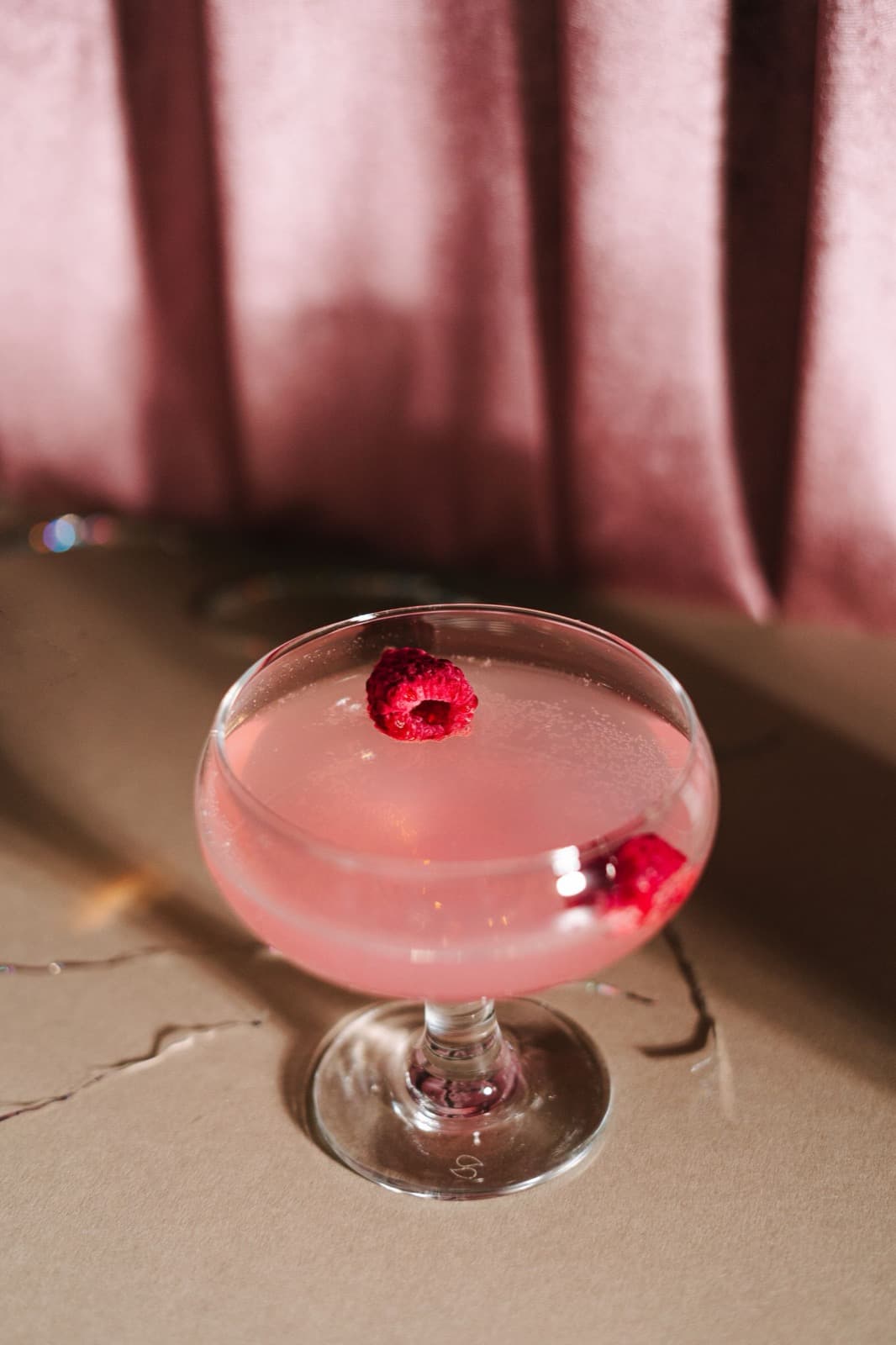 A pink cosmo in a coupe garnished with raspberries, on a warm stone surface against a blush curtain