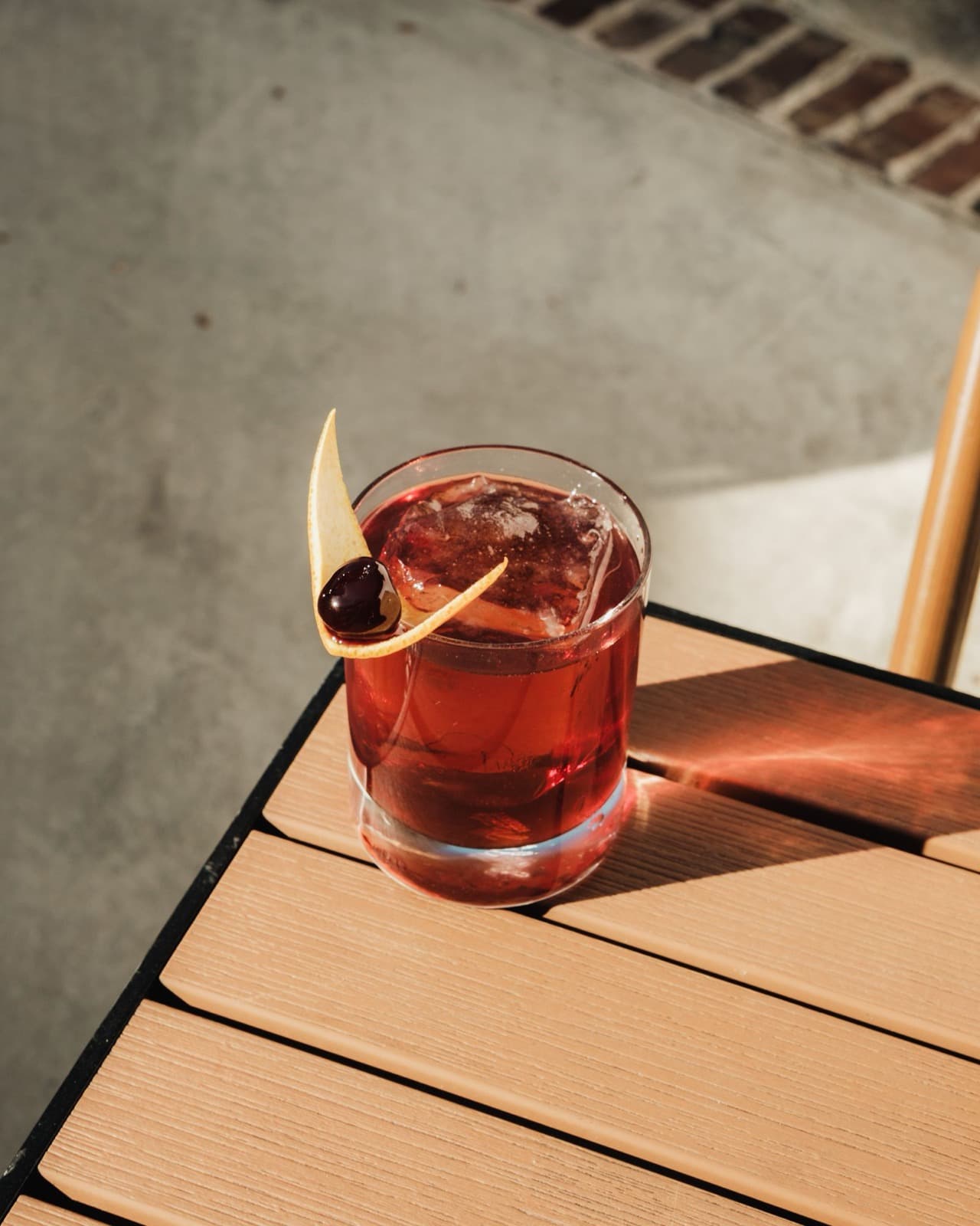 A deep red drink on ice in a rocks glass with an orange peel and cherry, on a sunlit wooden table