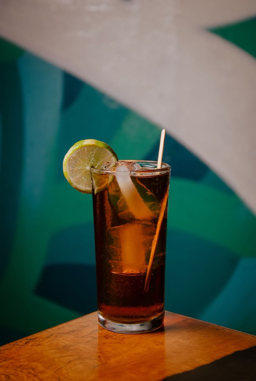 A highball of amber ginger drink over ice with a lime wheel and straw, on a wooden ledge against a painted wall