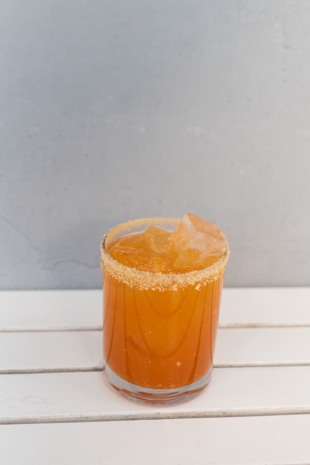 An orange spicy margarita on ice in a tumbler with a spiced salt rim, on a white slatted table
