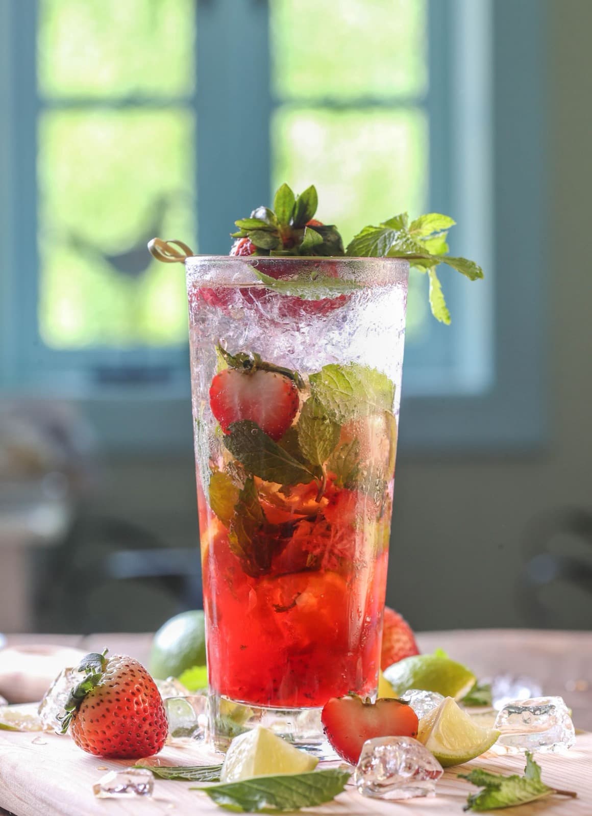 A tall strawberry mojito with muddled strawberries, mint and crushed ice, beside fresh strawberries and lime on a pale board by a window