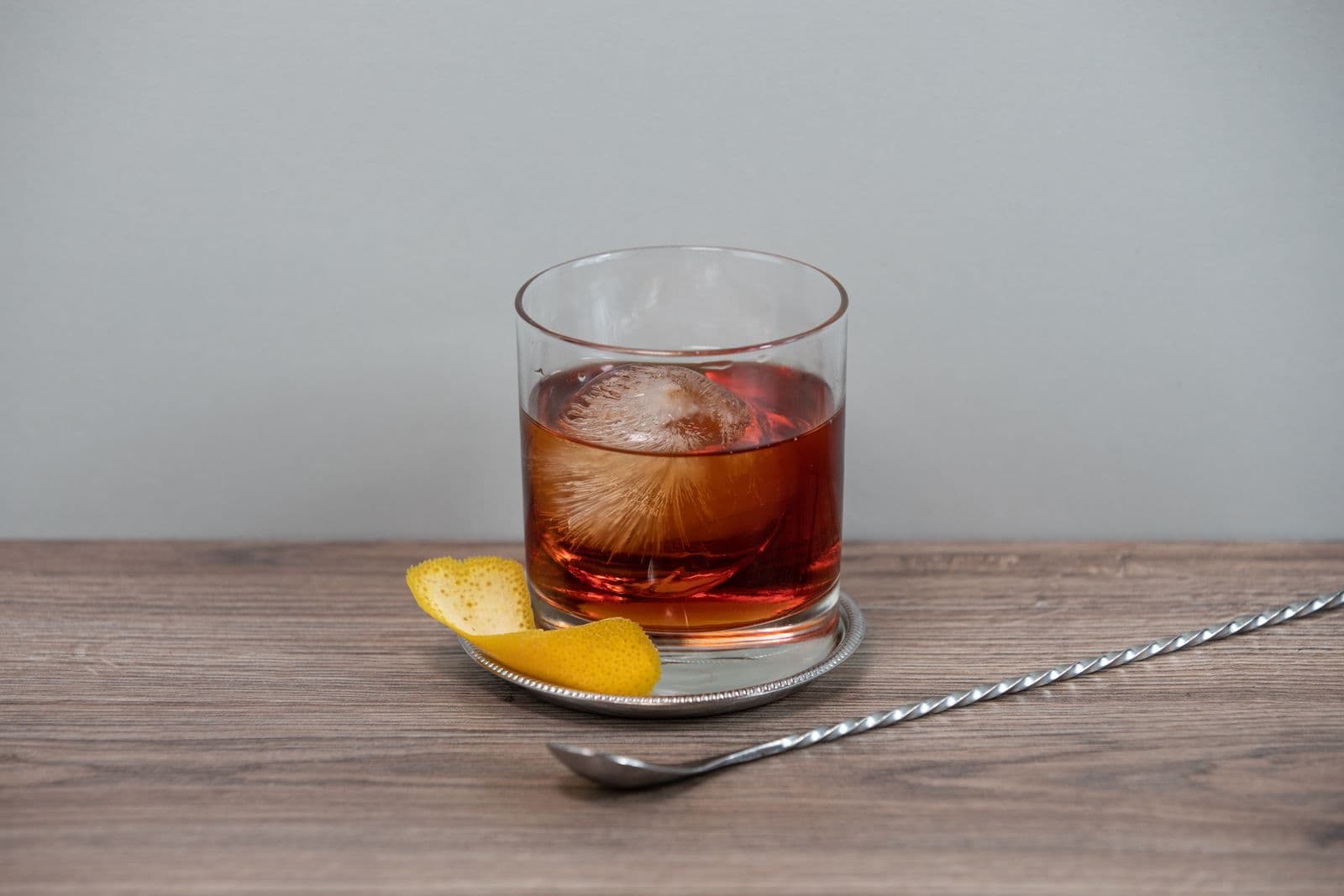 An amber drink over a large ice cube in a rocks glass with an orange peel on the saucer, on a light wooden counter against a grey wall