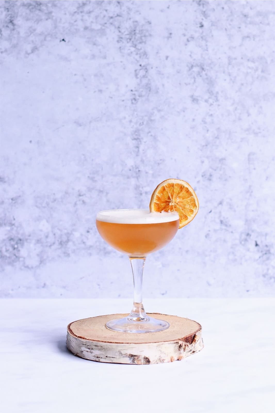 A whiskey sour with a foam top and a dried orange wheel in a coupe, on a birch slice against pale concrete