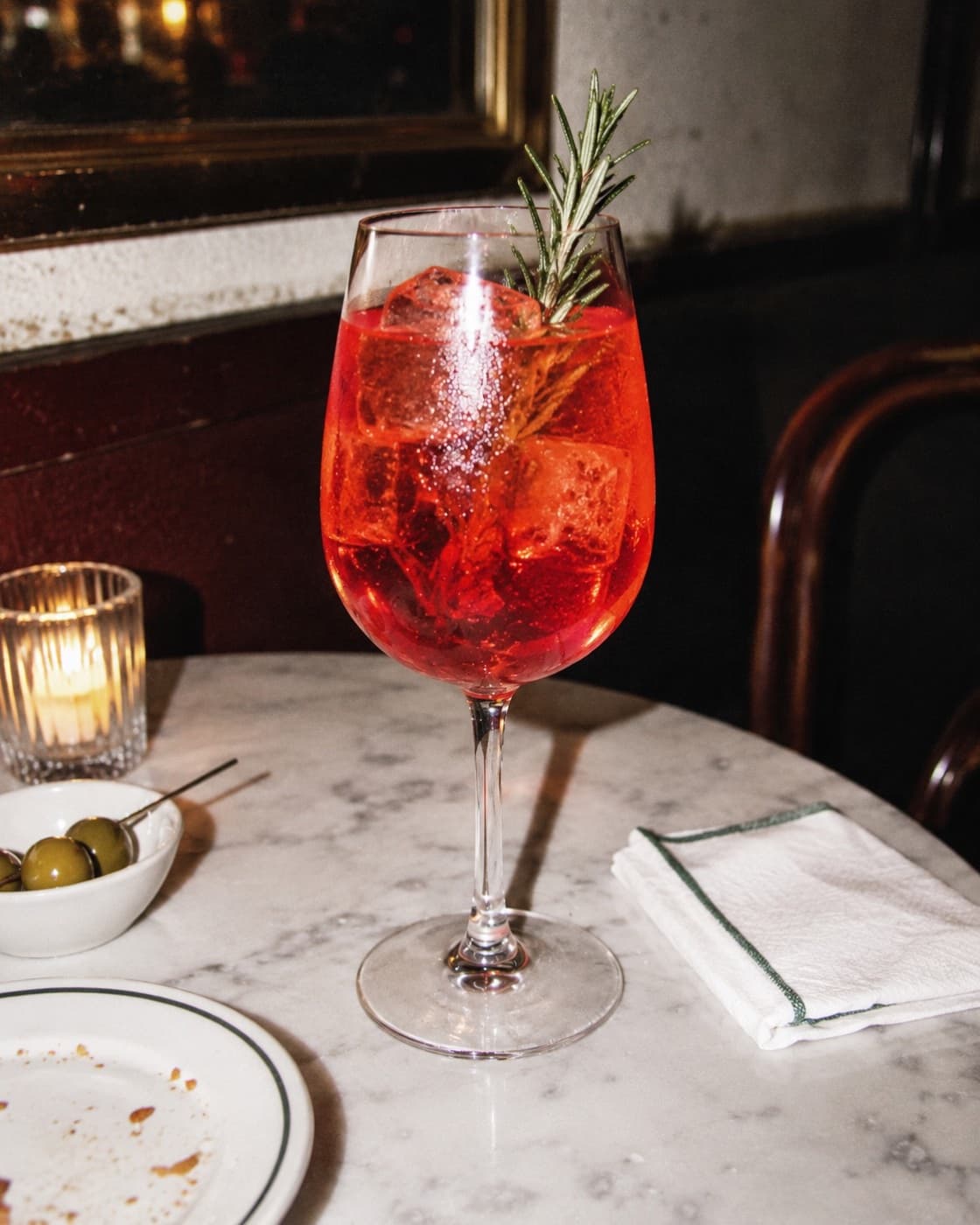 Looking down into a red-orange spritz on ice with a blood orange slice and a rosemary sprig, on pale pink marble