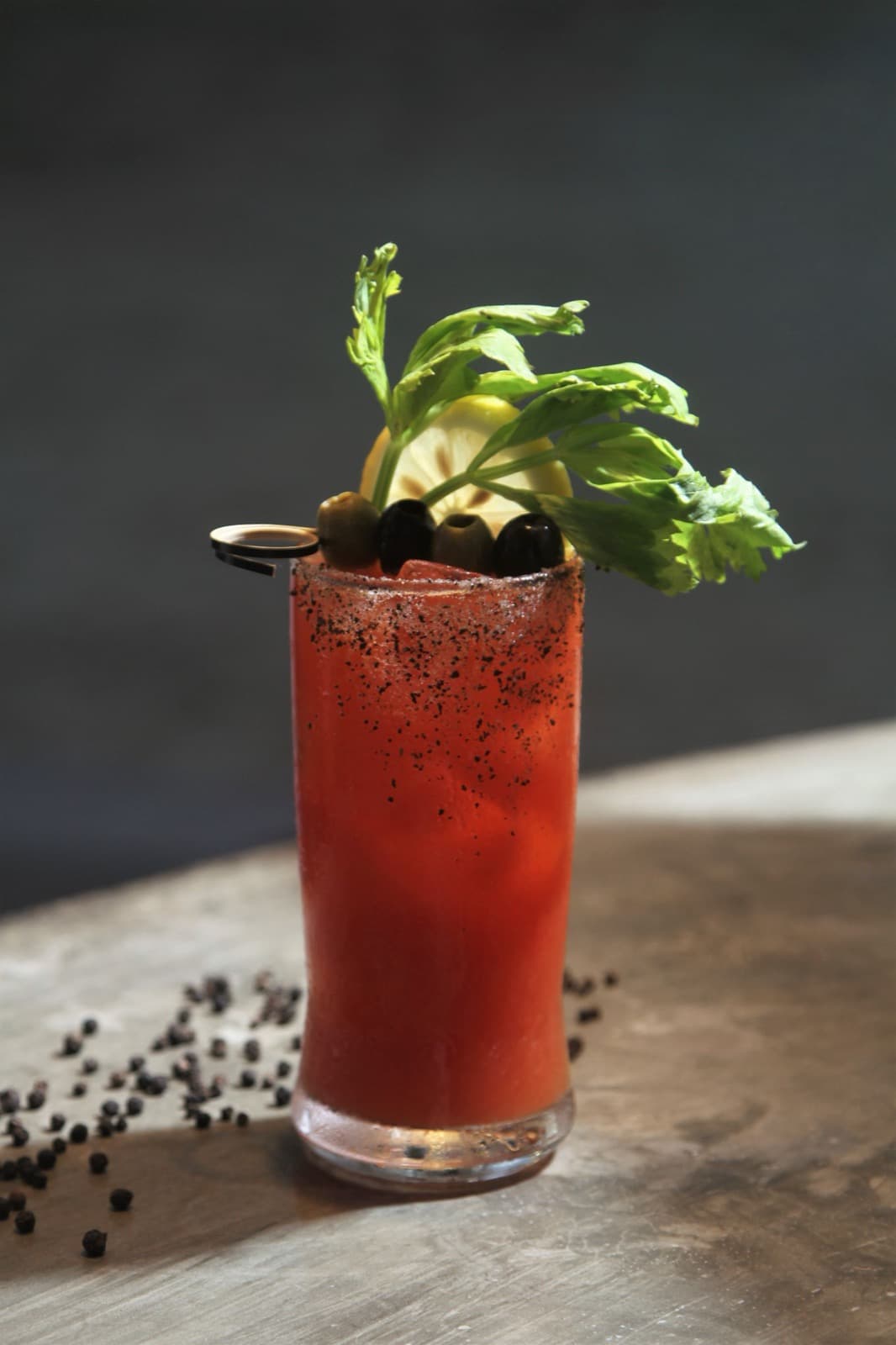 A tall Bloody Mary with a pepper rim, garnished with a celery top and a pick of olives, on a stone surface scattered with peppercorns