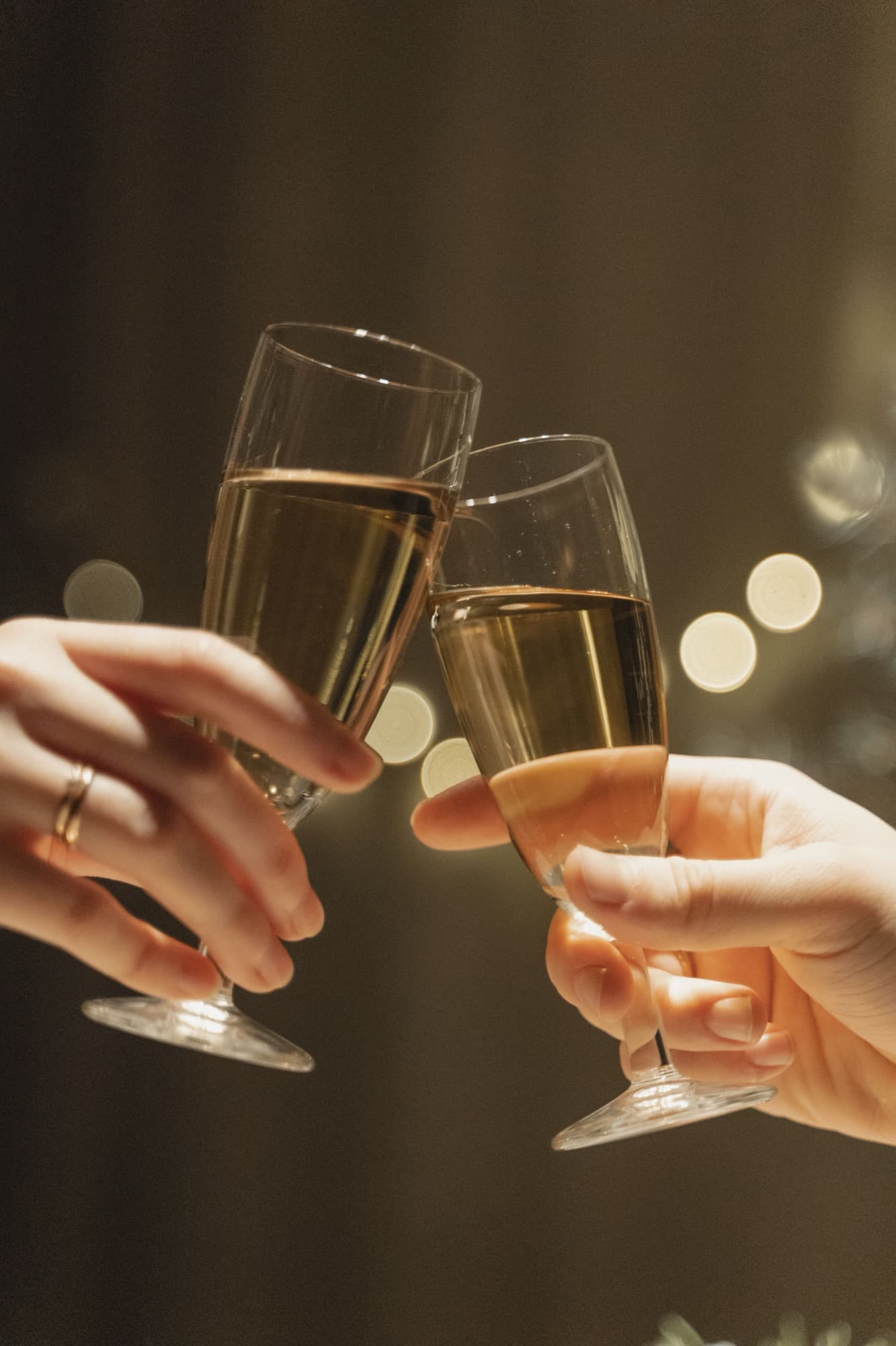 Two hands clinking sparkling glasses against a warm, dark background with soft bokeh lights