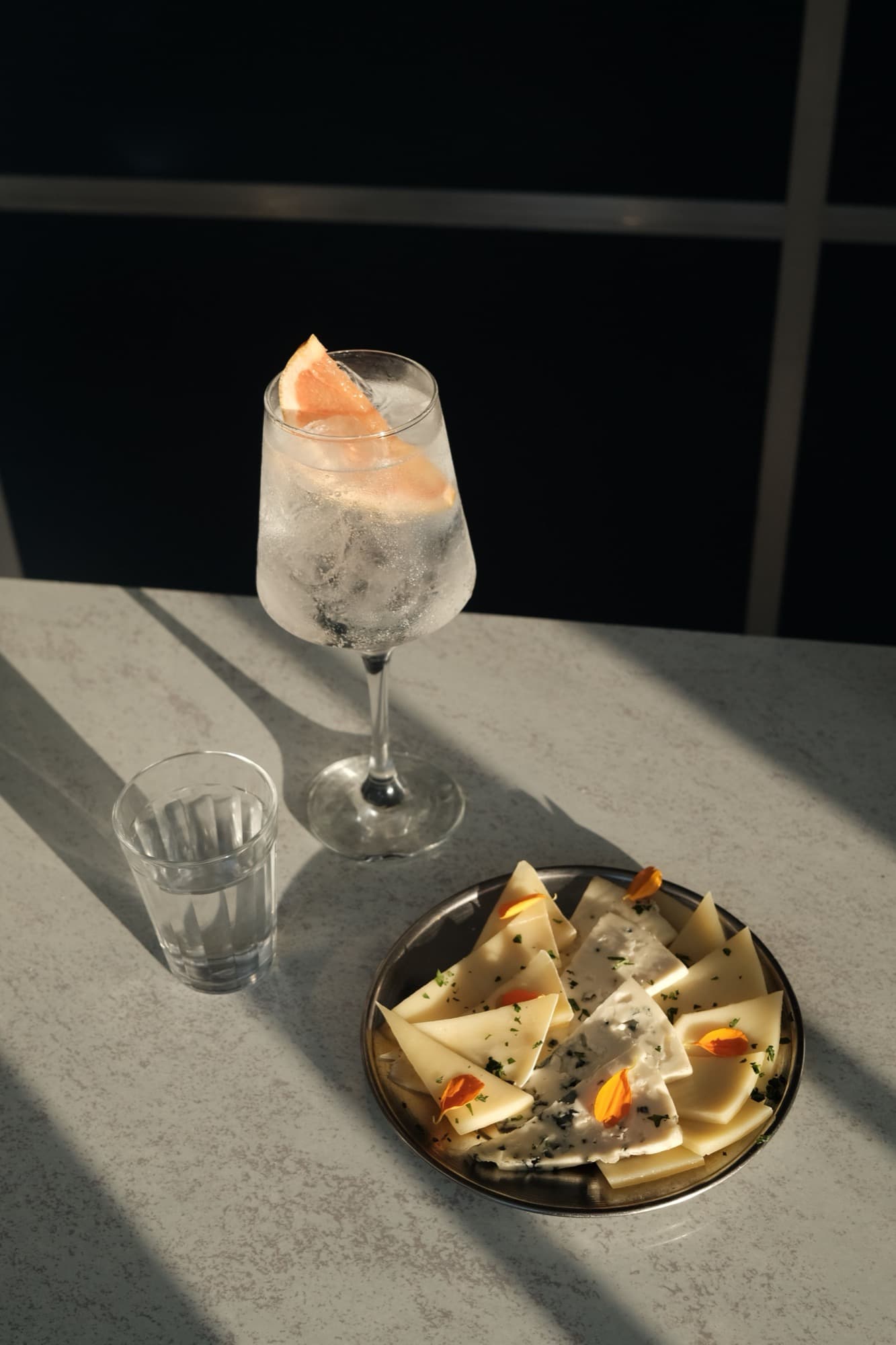 A spritz with a grapefruit wedge, a glass of water and a plate of cheese on a stone table in low afternoon sun