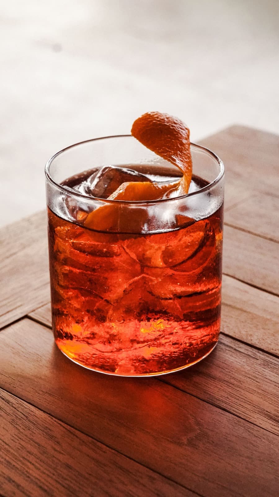 A deep red-orange negroni-style drink on ice with an orange peel twist in a short tumbler, on a wooden table against a pale wall