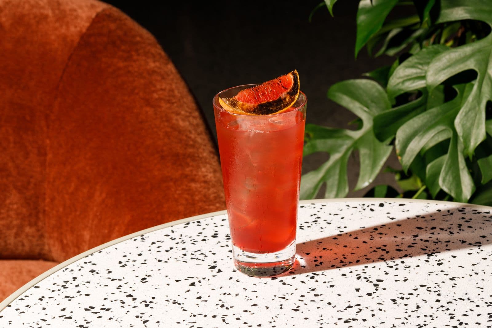 A tall red drink in a highball glass with a citrus wedge, on a speckled terrazzo table beside green leaves