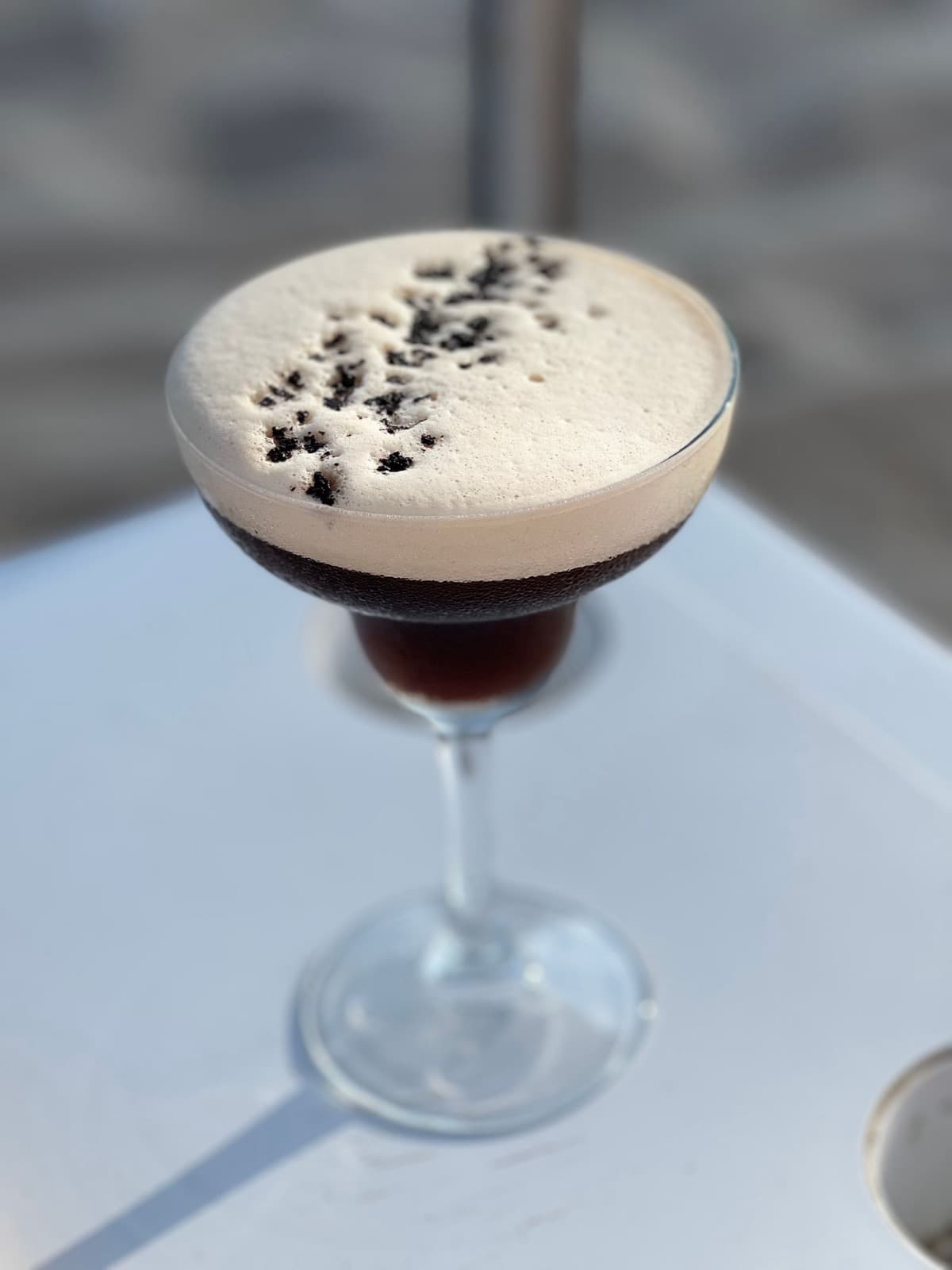 An espresso martini with a tall pale crema scattered with coffee beans, on a white table in daylight