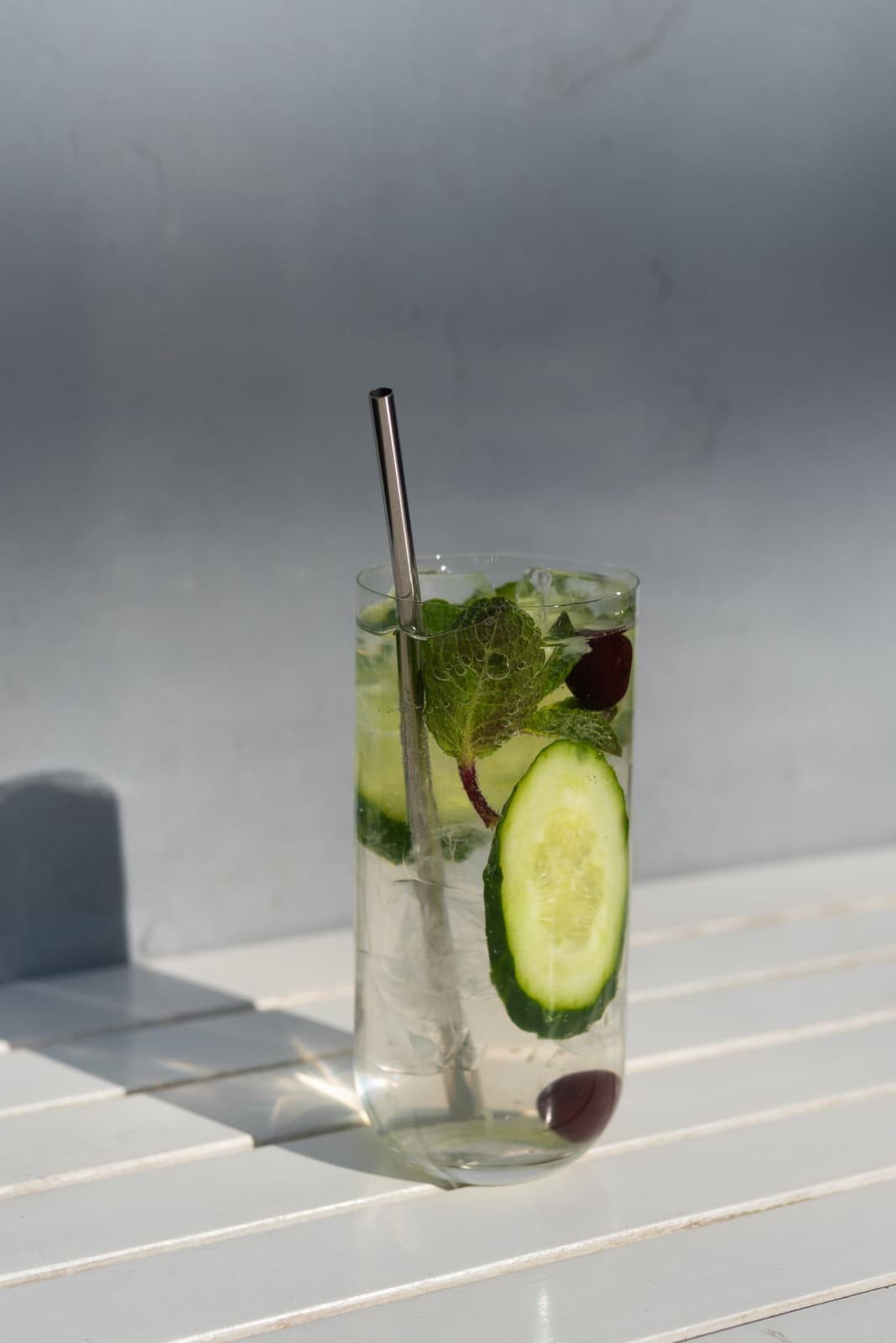 A gin and tonic in a tall glass with cucumber, mint and a metal straw on a white slatted table