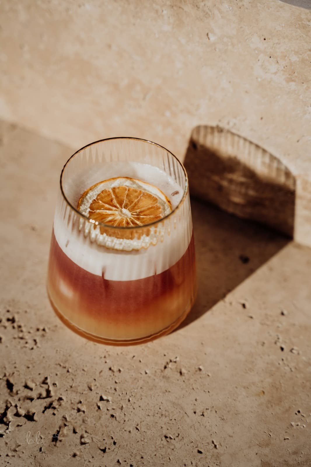 An amber sour with a pale foam cap and a dried citrus wheel in a ribbed rocks glass, on warm travertine in soft sunlight