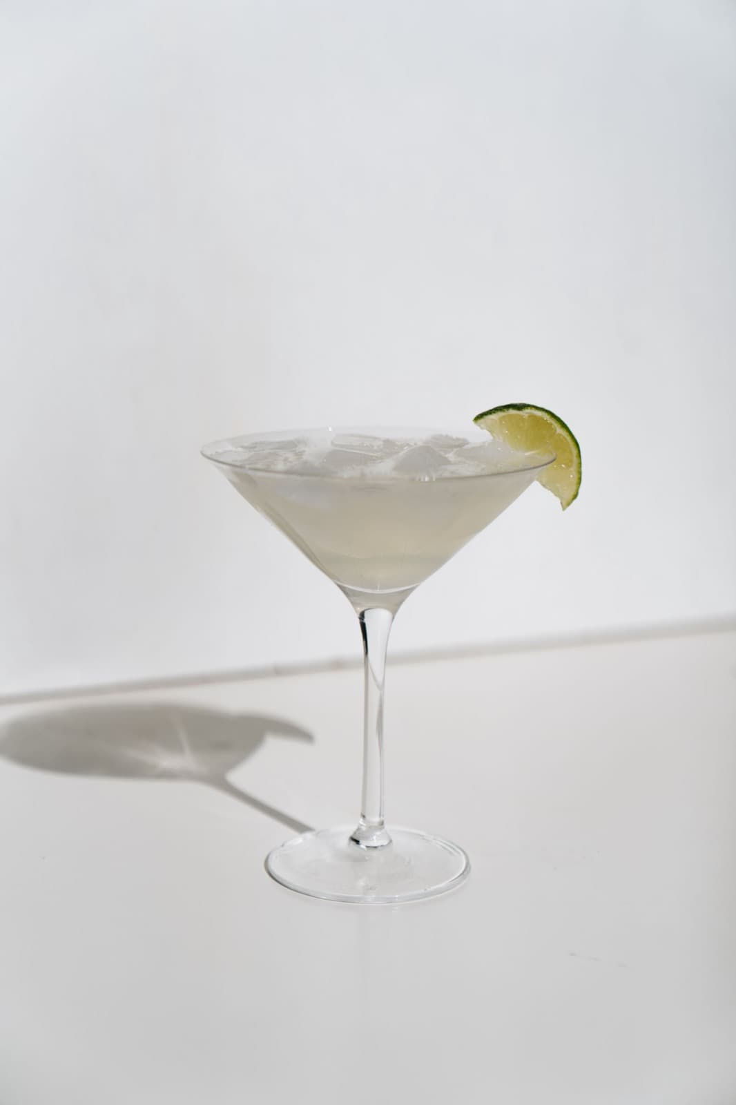 A margarita over crushed ice in a stemmed cocktail glass with a lime wedge on the rim, on a white table in sunlight