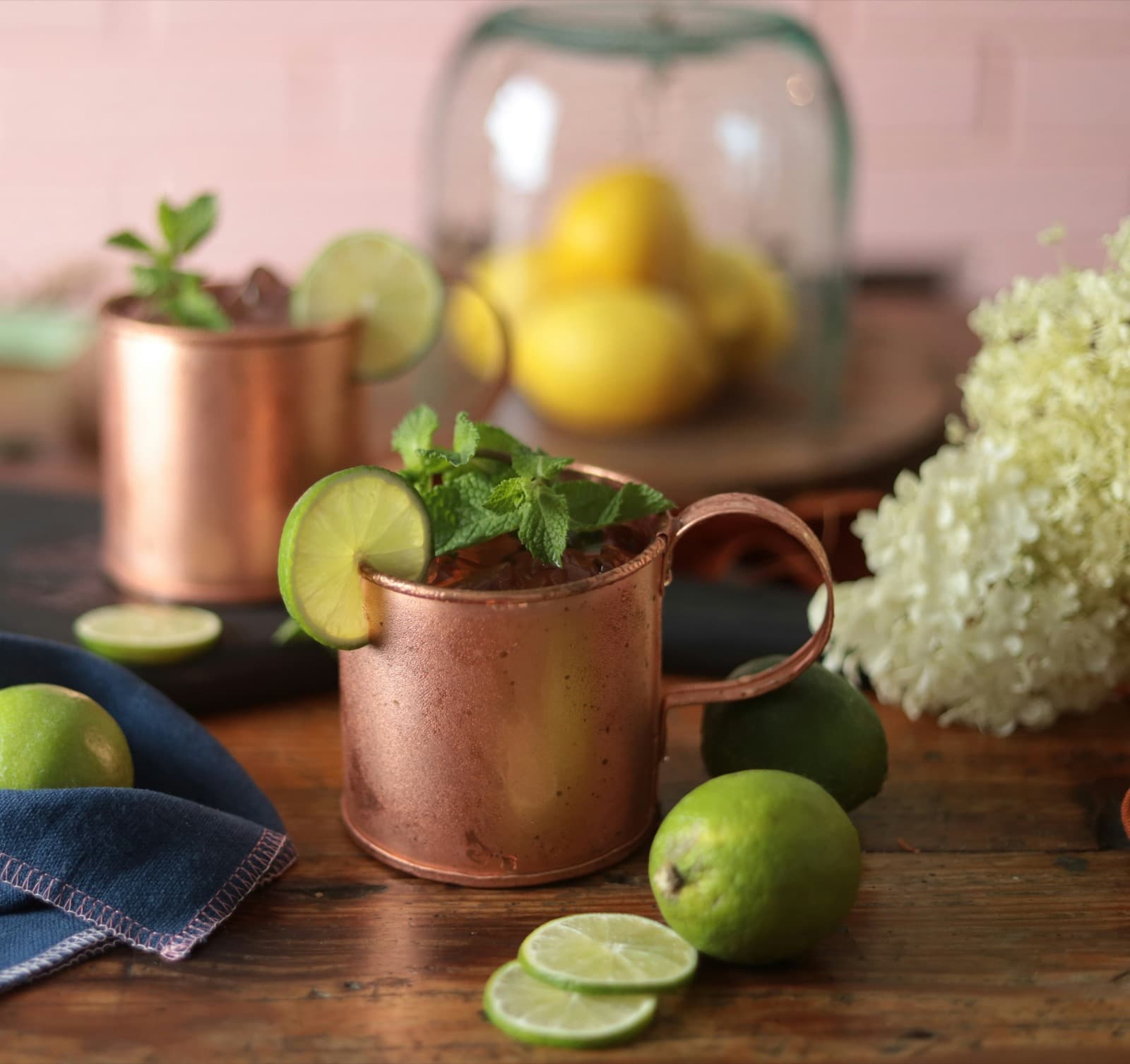Two copper mugs of iced ginger mule with mint and lime wheels, surrounded by fresh limes and white hydrangeas on a wooden table