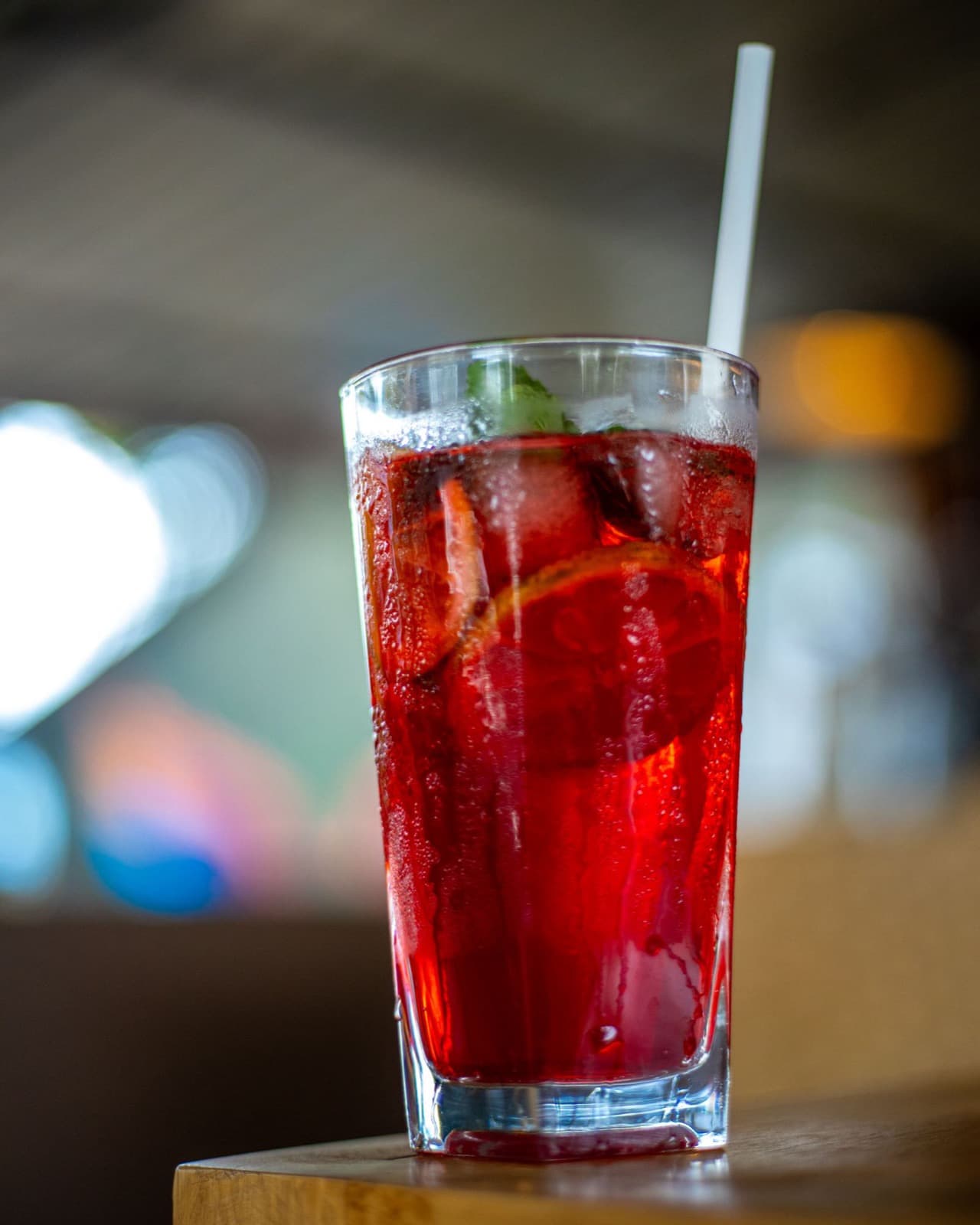 A bright red drink over ice in a tall highball glass with a citrus slice and a white straw