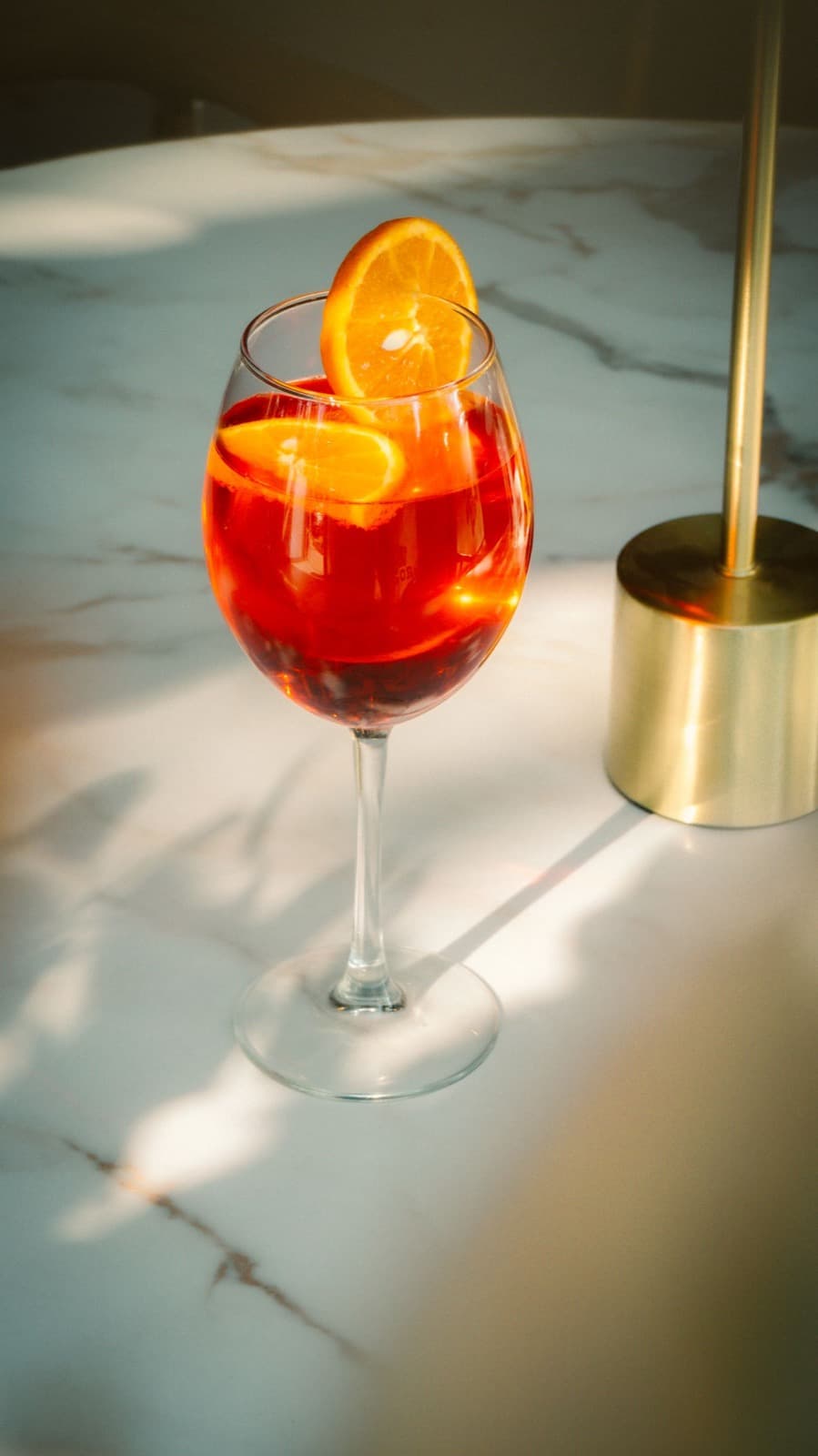 A red-orange spritz in a wine glass with orange slices, on a sunlit white marble table