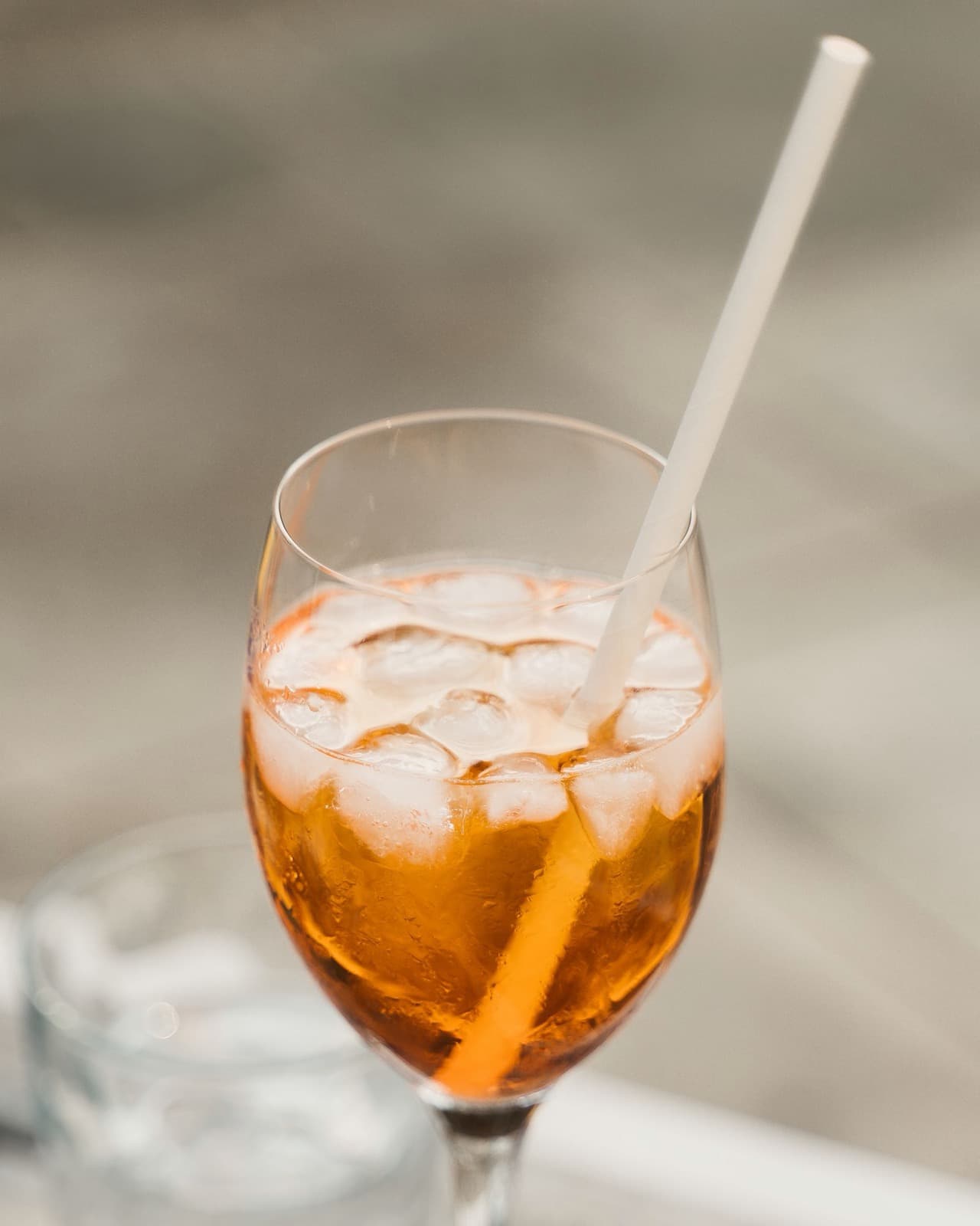 An orange spritz over ice in a wine glass with a white paper straw, against a soft pale background