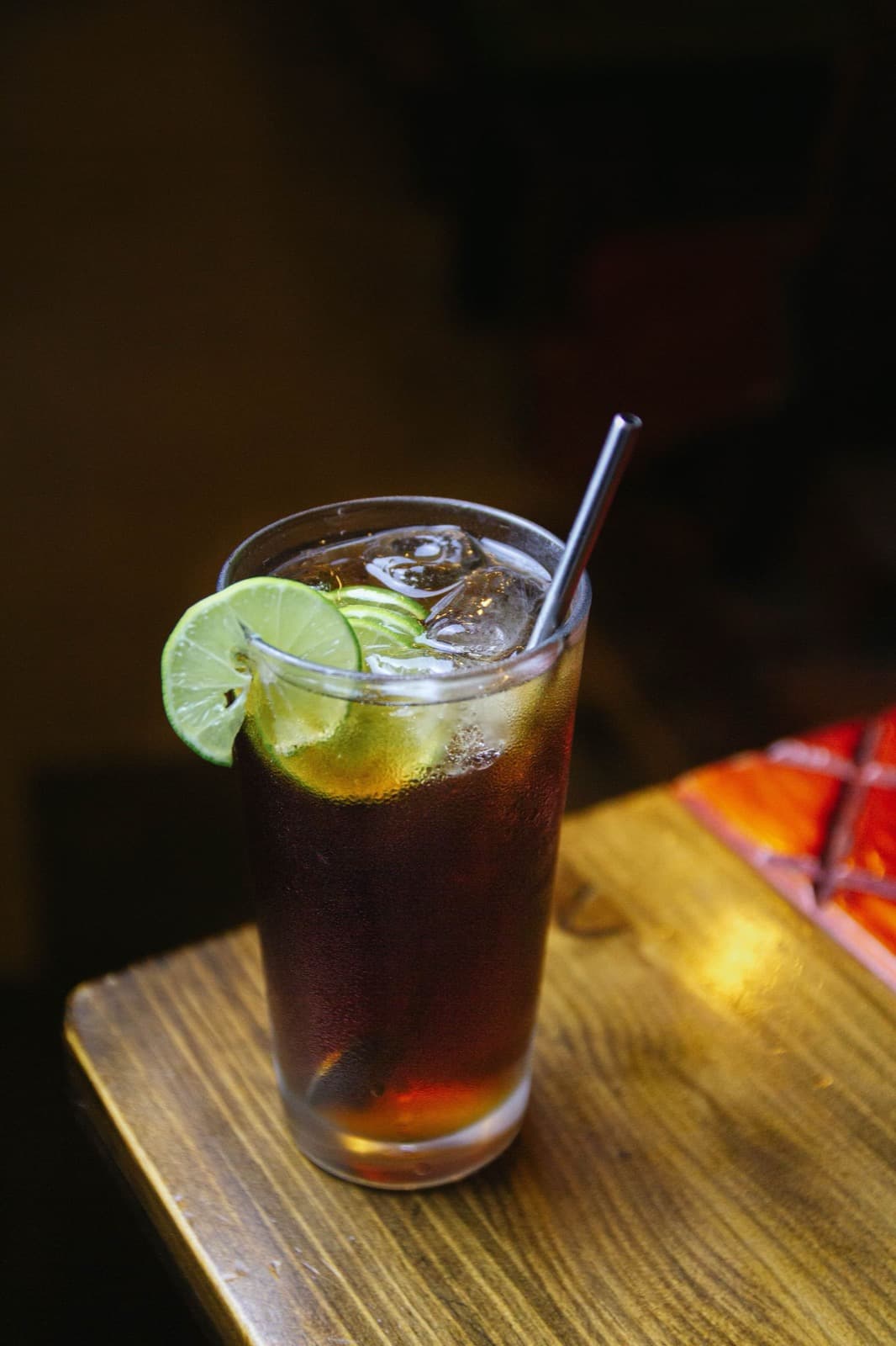 A tall glass of dark amber ginger drink over ice with lime wheels and a straw, on a warm wooden table