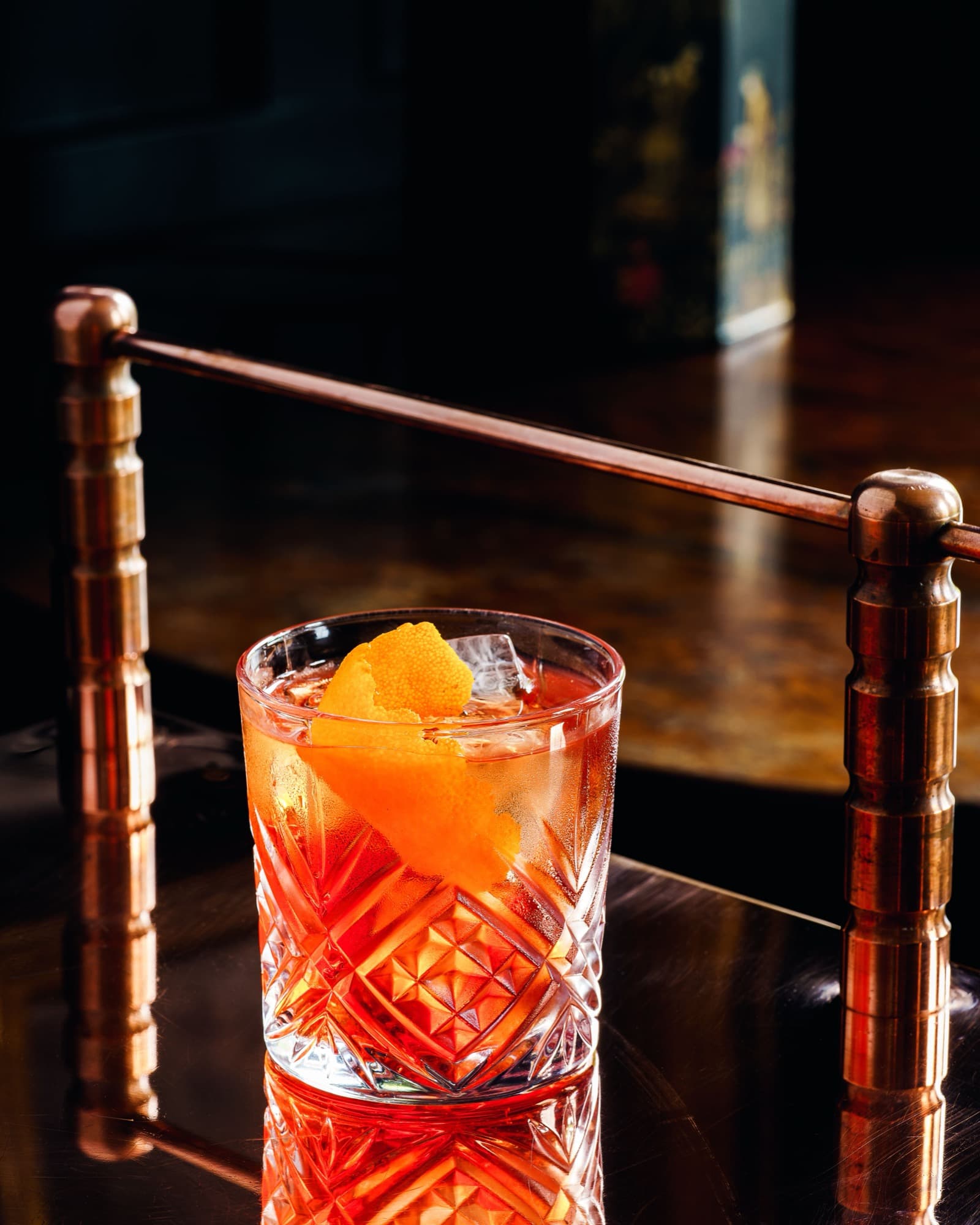 A Negroni-style drink in a cut-crystal glass with an orange twist and ice, on a dark polished bar behind a brass rail